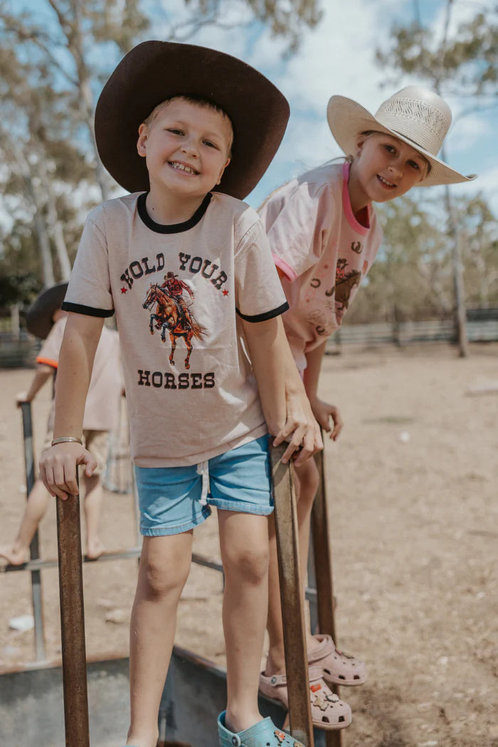 Milky Hold Your Horses Tee. Two children in cowboy hats and shirts standing on a wooden fence with trees in the background.