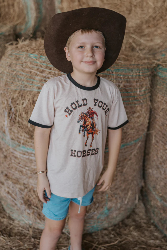 Milky Hold Your Horses Tee. Young boy wearing a cowboy hat and 'Hold Your Horses' t-shirt standing in front of hay bales.