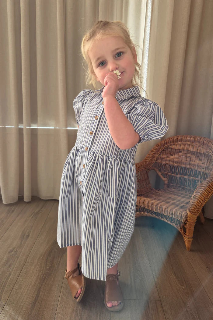 Milky Girl's Marine Stripe Dress. Young girl in a striped dress standing indoors with curtains and a chair in the background.