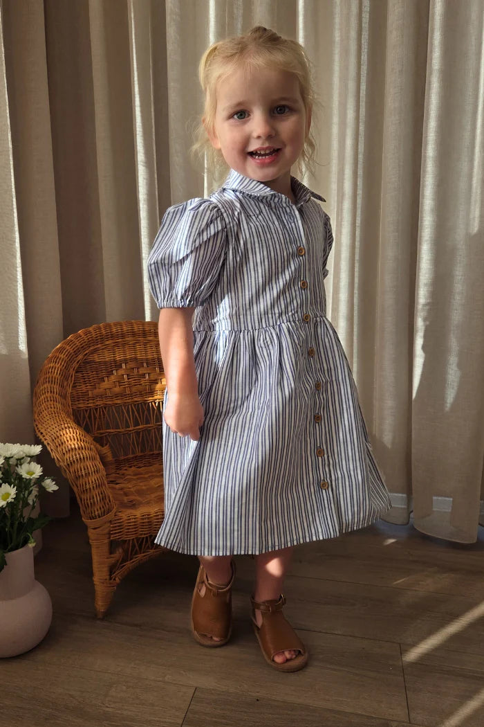 Milky Girl's Marine Stripe Dress. Young girl in a striped dress standing in a room with curtains and a wicker chair.