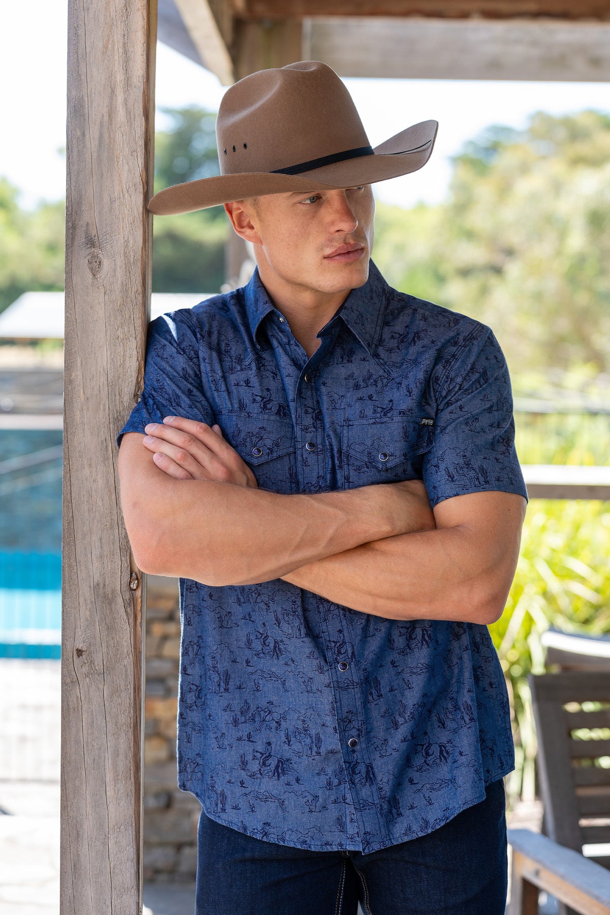 Pure Western Men's Travis S/S Western Shirt. Man wearing a blue patterned shirt and brown cowboy hat standing with arms crossed outdoors.