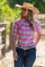 Pure Western Women's Leni S/S Western Shirt. Woman wearing a pink plaid shirt, blue jeans, and a cowboy hat outdoors.