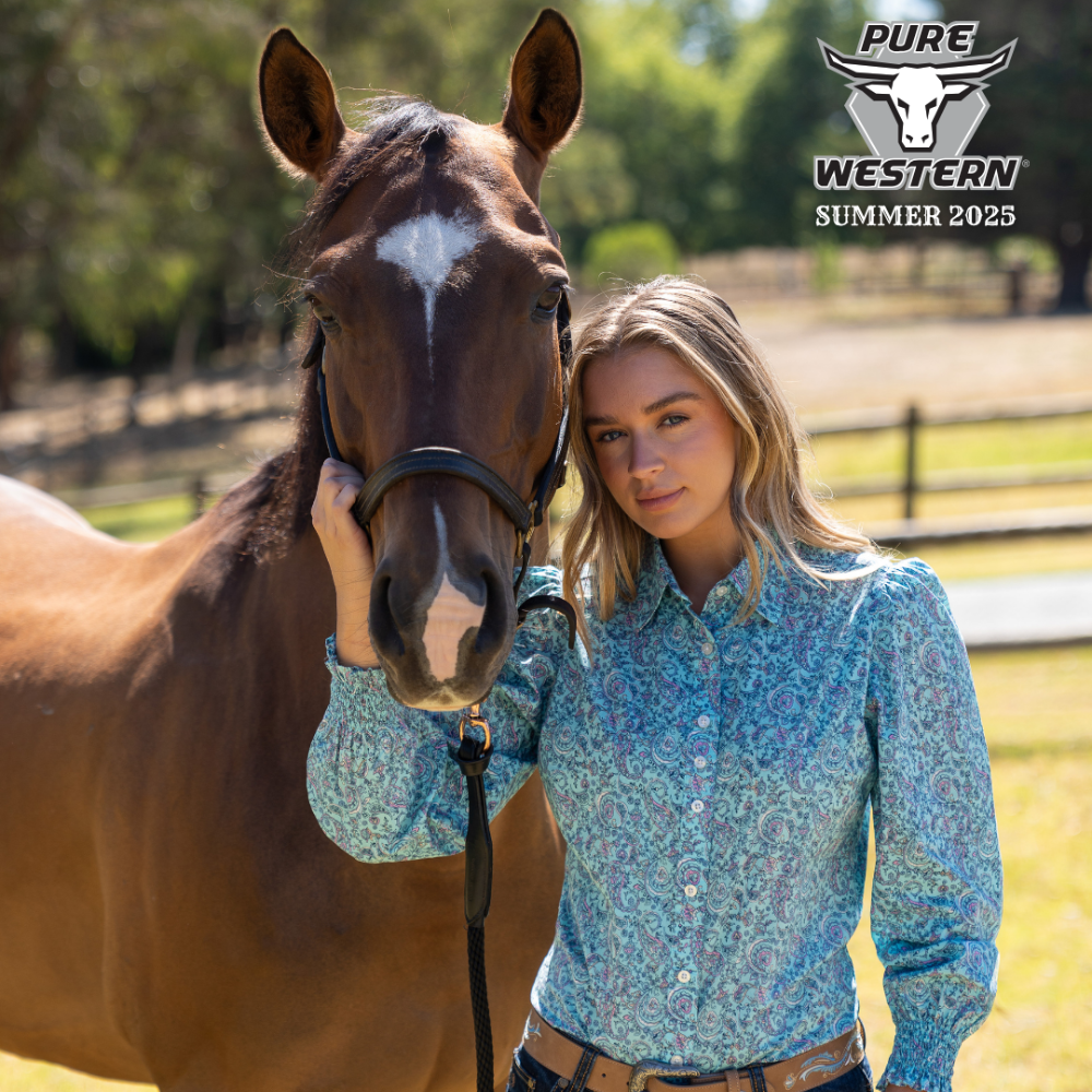 Pure Western Women's Riley L/S Western Shirt. Woman in a blue patterned shirt standing next to a horse with 'PURE Western Summer 2025' logo in the corner.