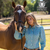 Pure Western Women's Riley L/S Western Shirt. Woman in a blue patterned shirt standing next to a horse with 'PURE Western Summer 2025' logo in the corner.