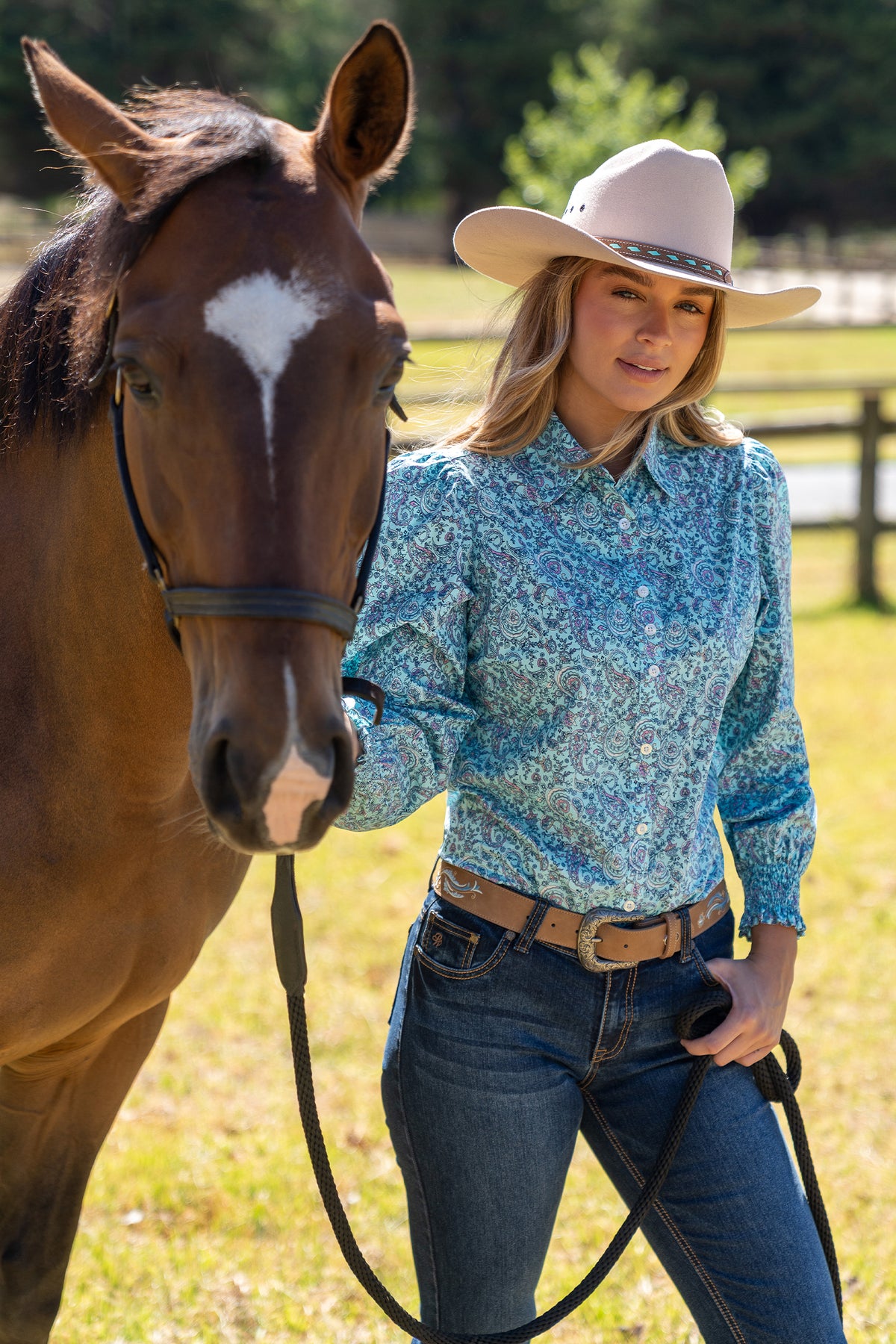 Pure Western Women's Riley L/S Western Shirt. Woman in a blue patterned shirt and cowboy hat standing next to a horse in a field.