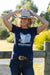 Pure Western Women's Sherry T-Shirt. Woman wearing a navy blue t-shirt with 'Western' design, holding a beige cowboy hat, standing outdoors near a wooden fence.
