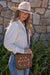 Pure Western Halley Western Handbag. Woman wearing a beige cowboy hat and holding a brown bag with decorative patterns against a stone wall.