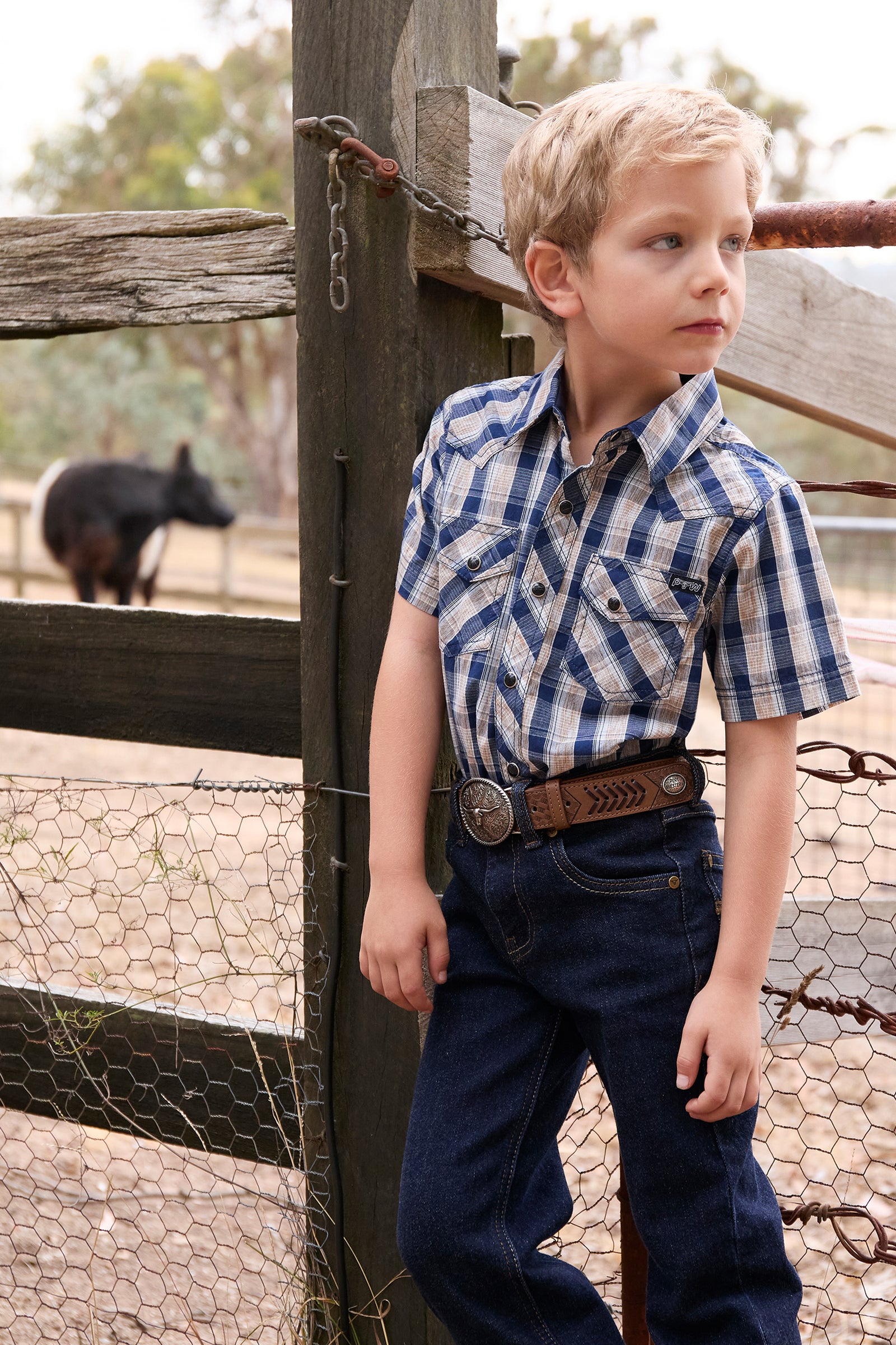 Pure Western Boy's Peter S/S Western Shirt. Young boy in a checkered shirt and jeans standing next to a wooden fence with a black animal in the background.