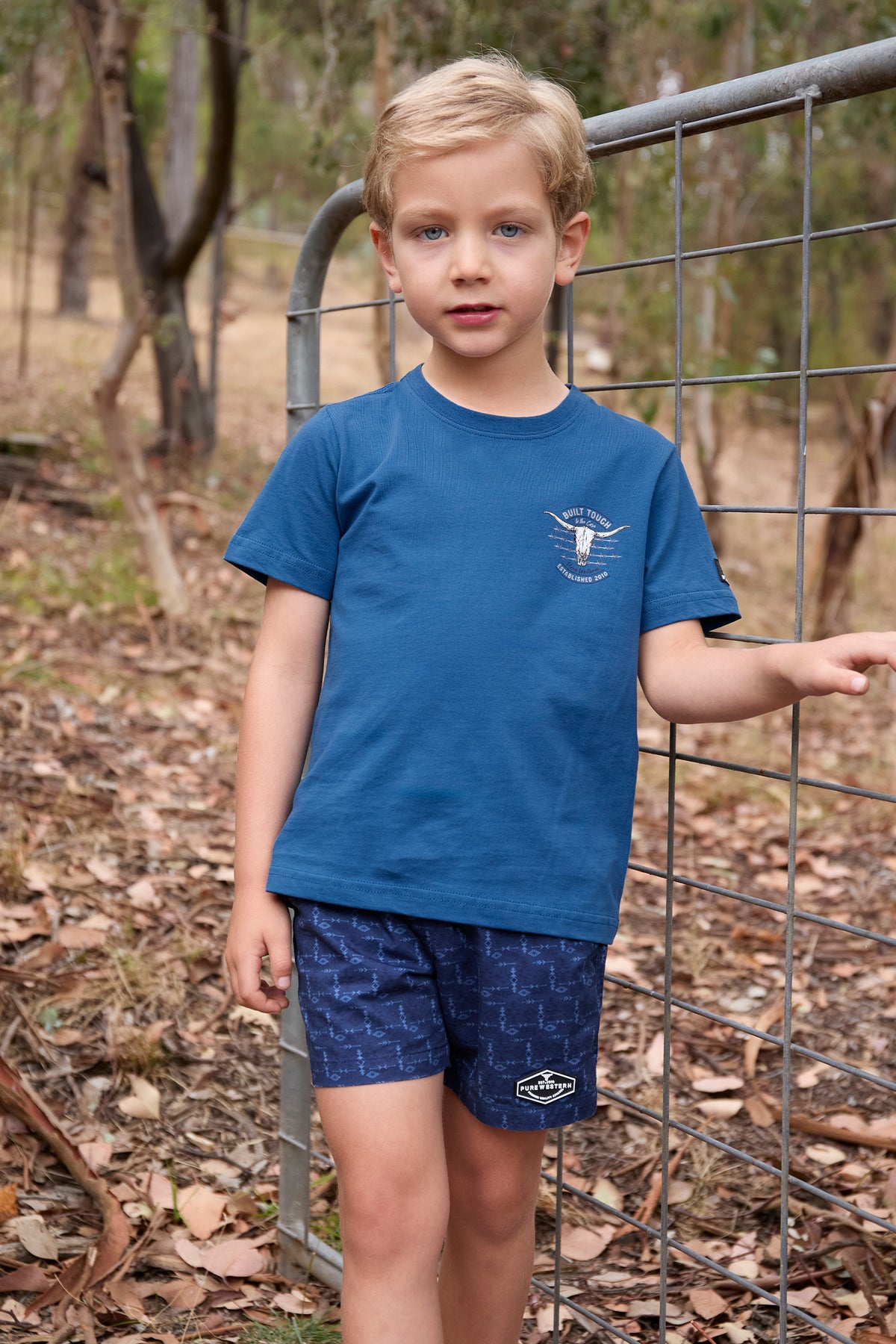 Pure Western Boy's Frank S/S T-Shirt. Young boy wearing a blue t-shirt and shorts standing near a wire fence outdoors.