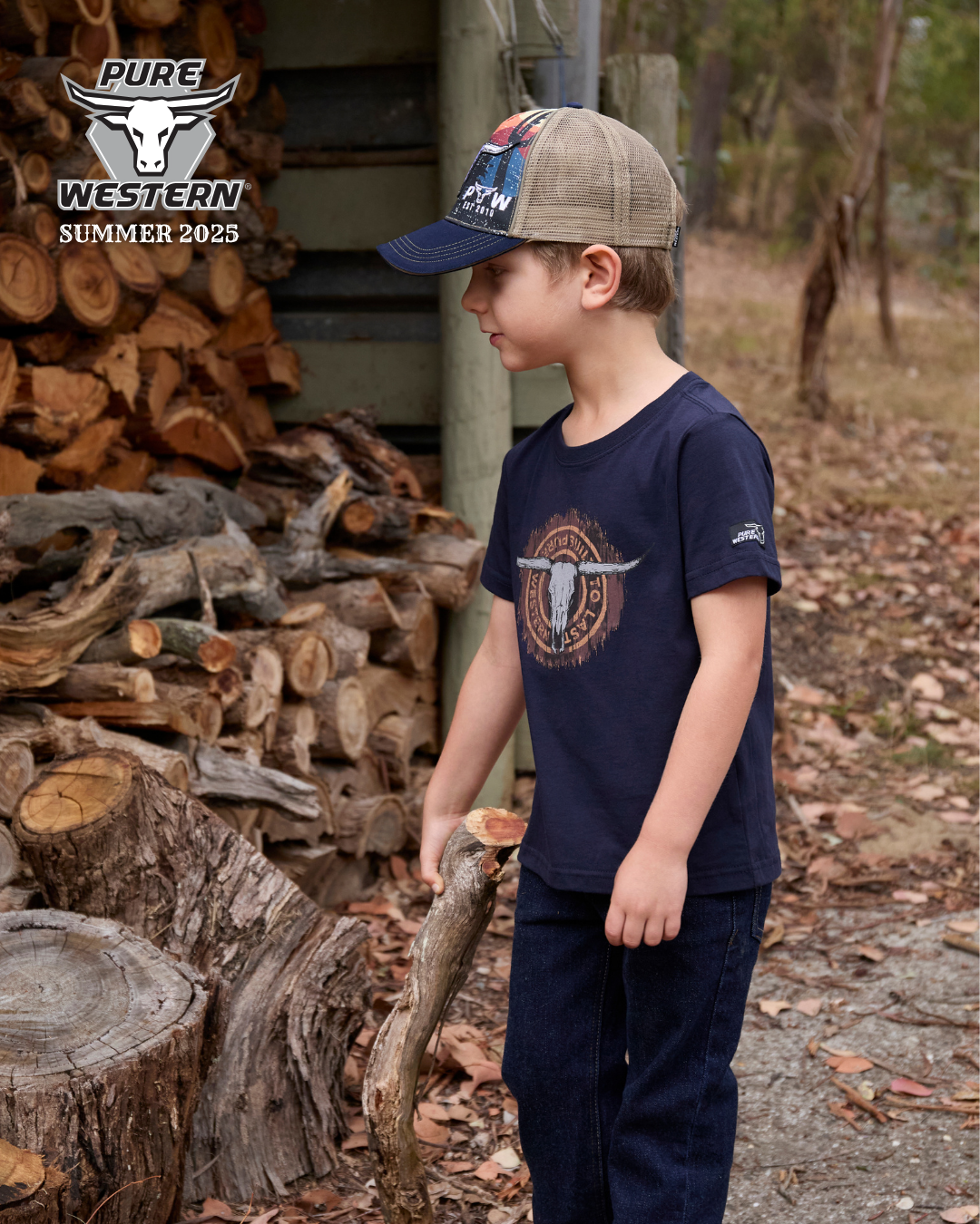 Pure Western Boy's Mason Trucker Cap. Child wearing a navy blue t-shirt with a logo, holding a stick, standing in front of stacked logs.