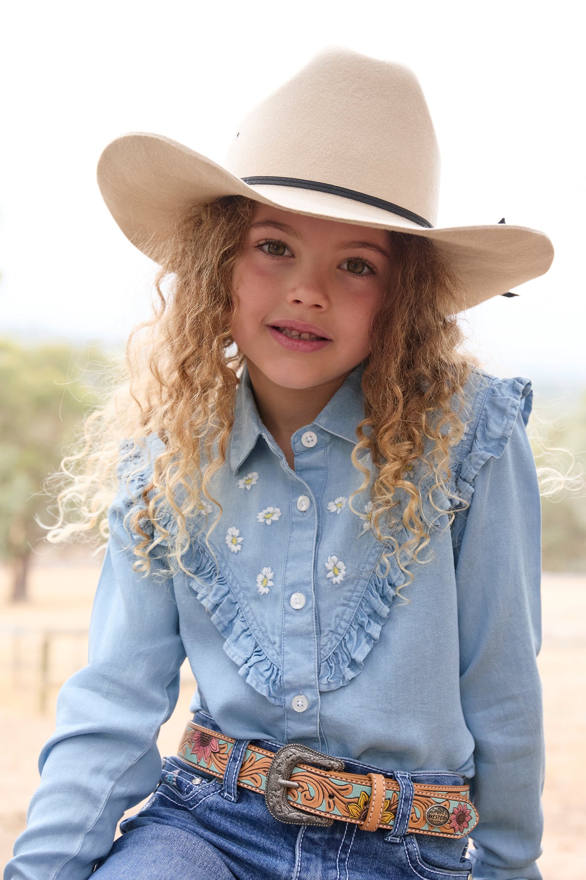 Pure Western Girl's Pheobe L/S Western Shirt. Young girl wearing a light blue embroidered shirt, cowboy hat, and colorful belt with a blurred outdoor background.
