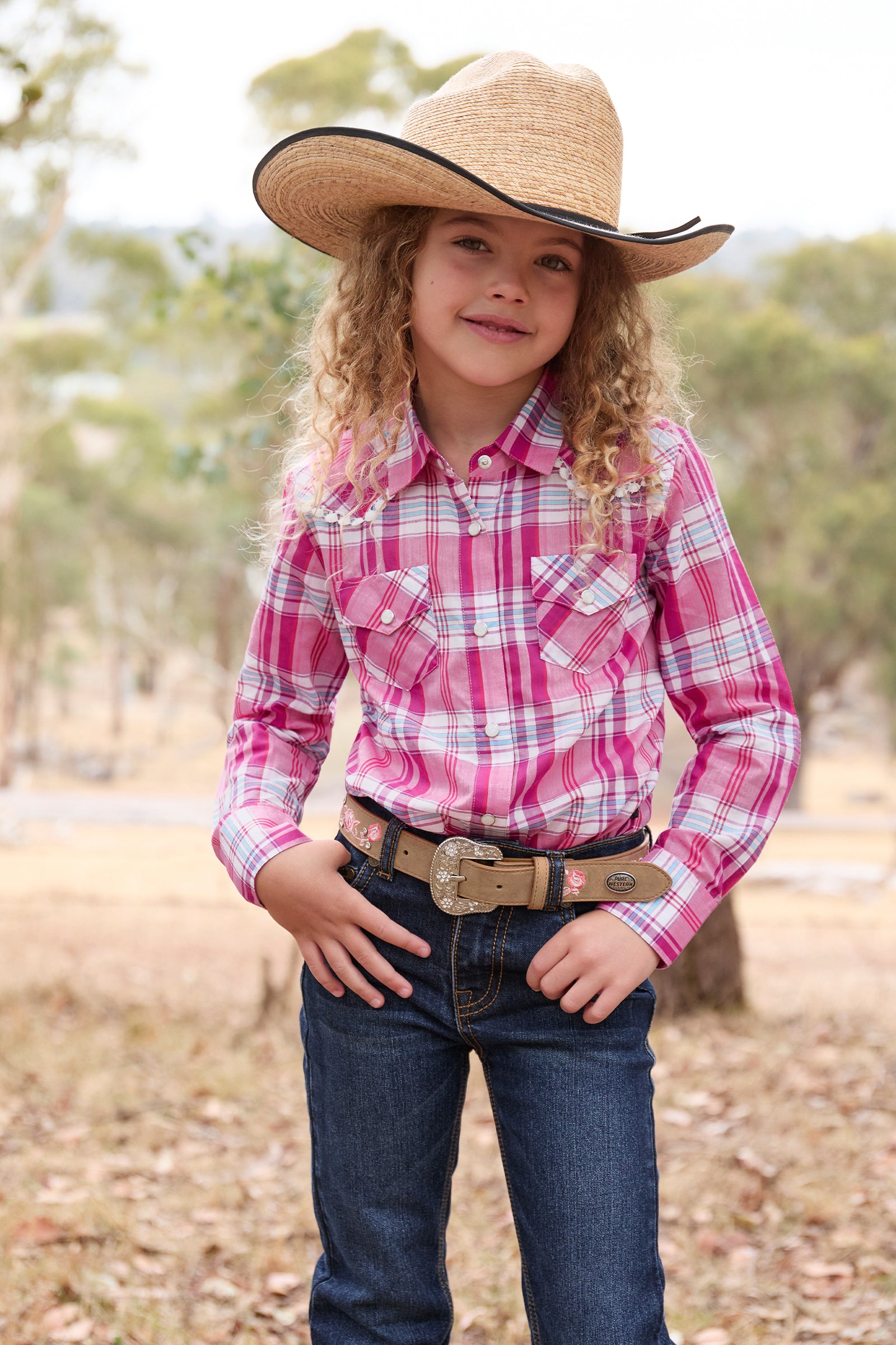 Pure Western Girl's Leni L/S Western Shirt. Child wearing a pink plaid shirt, blue jeans, and a straw cowboy hat outdoors.
