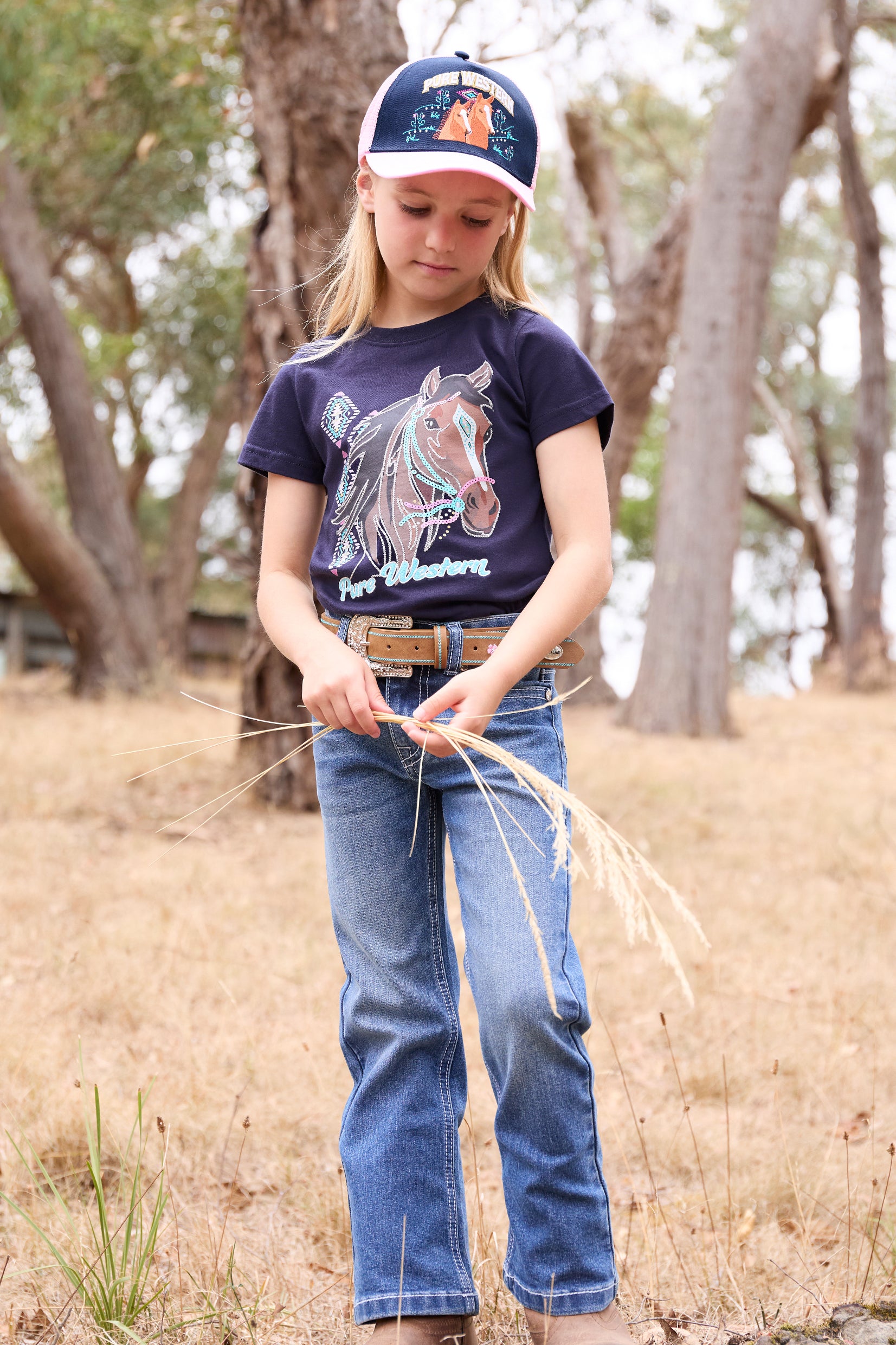 Pure Western Girl's Alesia Trucker Cap. Child in a natural setting wearing a navy t-shirt with a horse design and a cap.