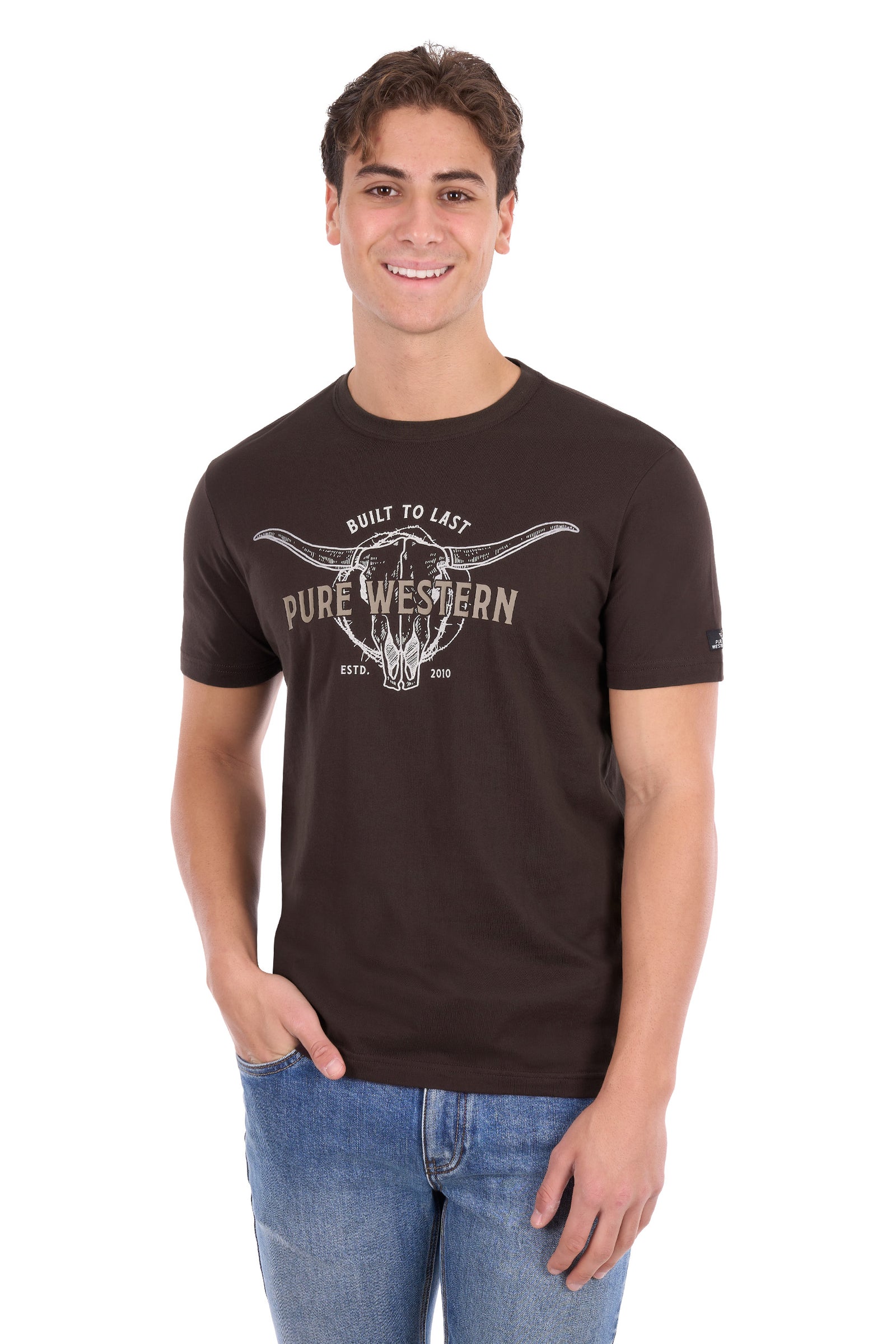A man wearing a dark brown short-sleeve t-shirt with a distressed white graphic that reads 'PURE WESTERN' and 'BUILT TO LAST' around a longhorn skull design, paired with blue jeans, against a plain white background.