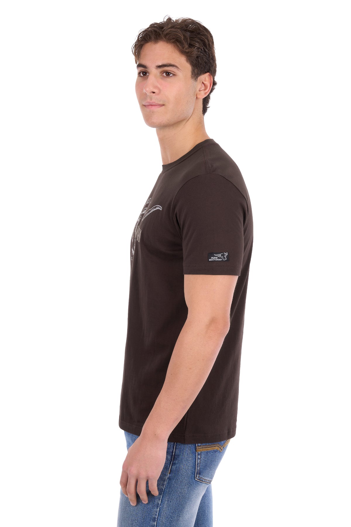 A man wearing a dark brown short-sleeve t-shirt with a small graphic on the left chest and a logo on the right sleeve, paired with blue jeans, shown in profile against a white background.