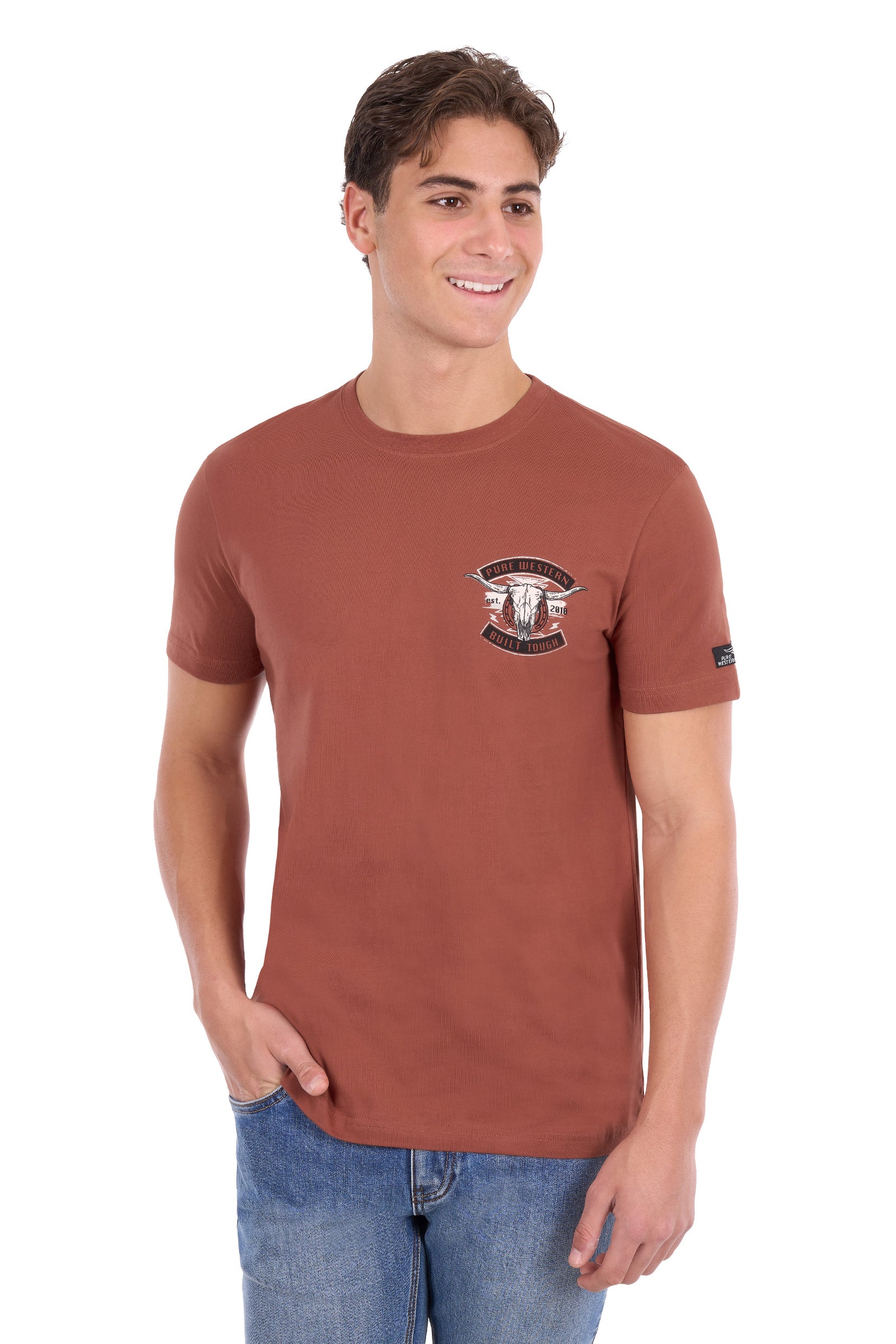A man wearing a rust-brown short-sleeve crewneck t-shirt with a small graphic on the left chest and a small black brand tag on the right sleeve, paired with blue jeans, against a plain white background.