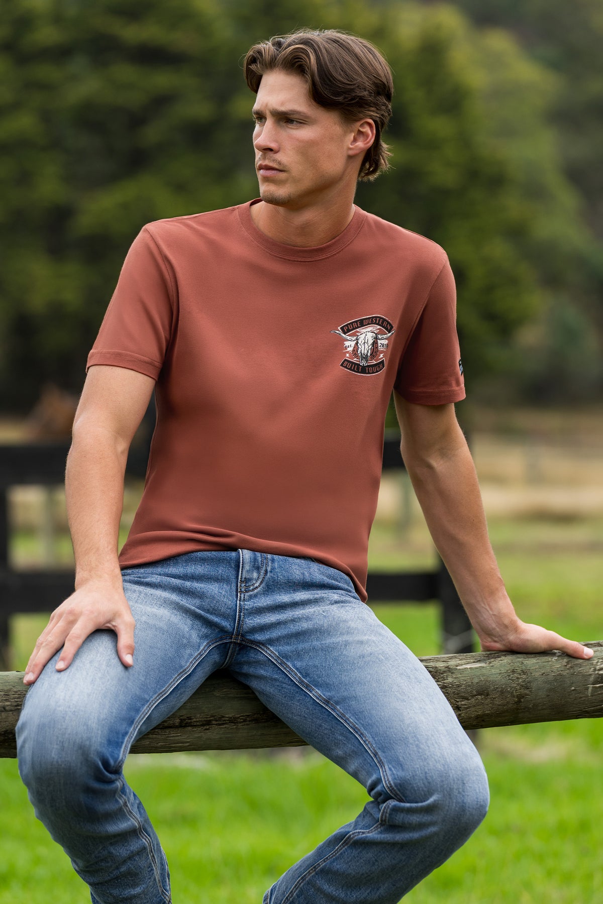 A man wearing a rust-brown short-sleeve t-shirt with a small embroidered logo on the left chest, paired with blue jeans, sitting on a wooden fence in an outdoor grassy field.