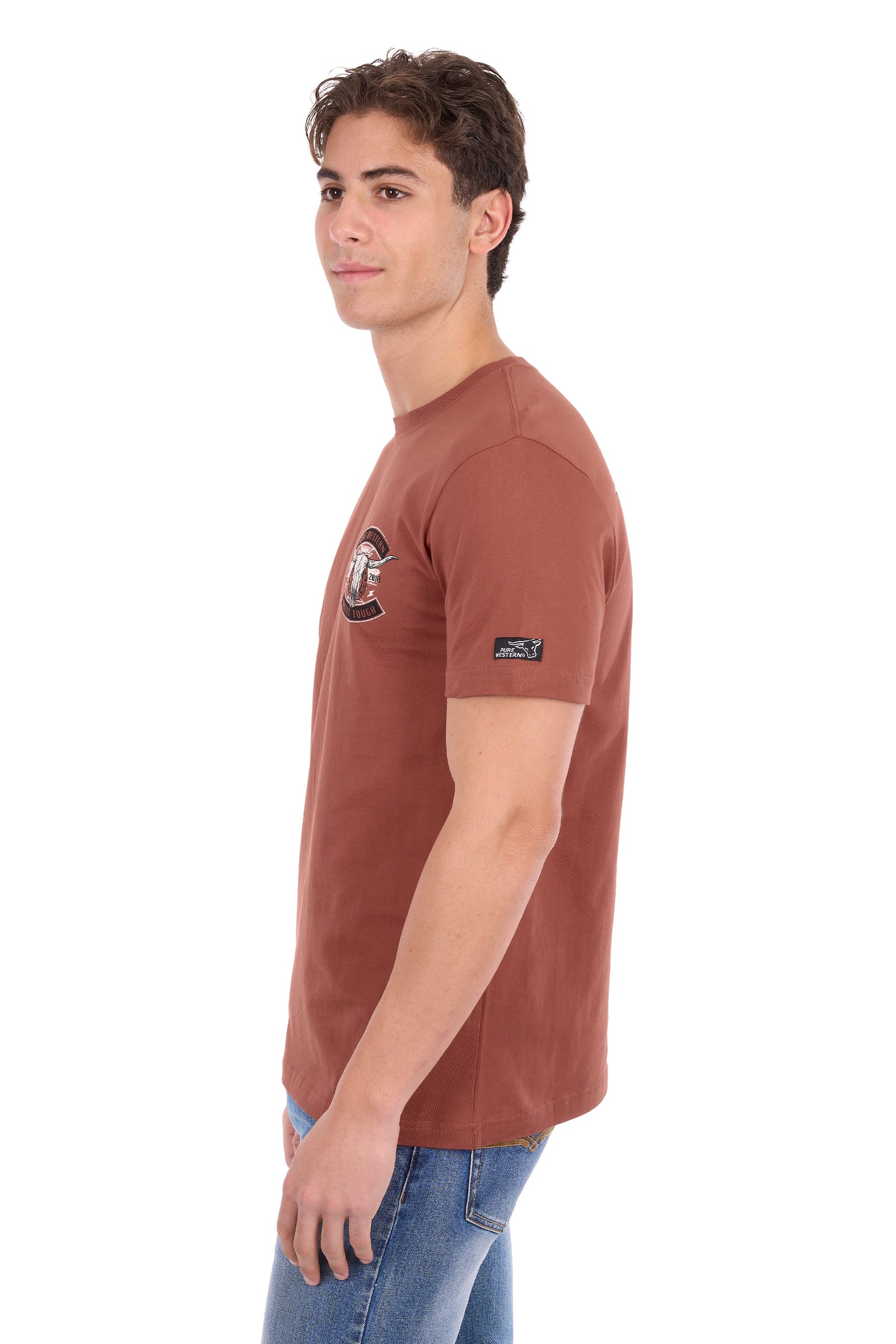 A man wearing a rust-brown short-sleeve t-shirt with a small graphic on the left chest and a brand patch on the sleeve, paired with blue jeans, photographed against a plain white background.