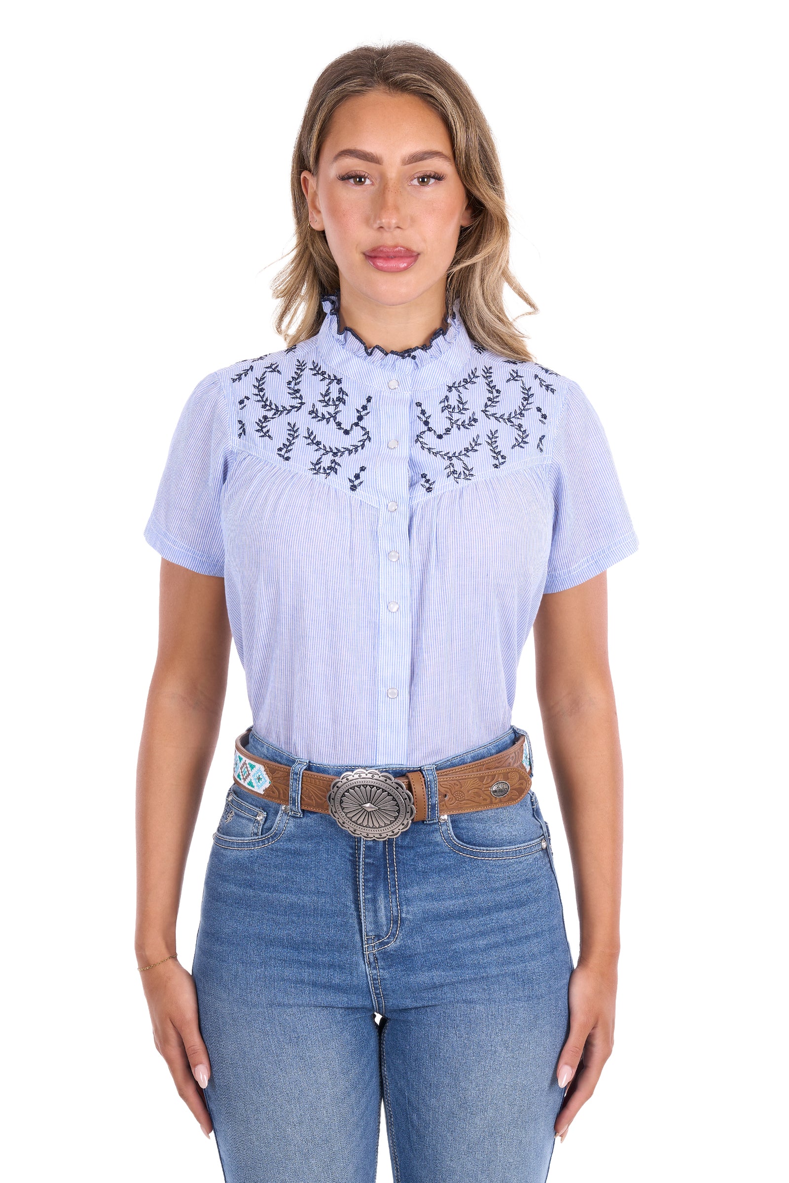 A woman modeling a light blue short-sleeve western-style blouse with black embroidered details on the chest, paired with blue jeans and a brown belt, against a plain white background.