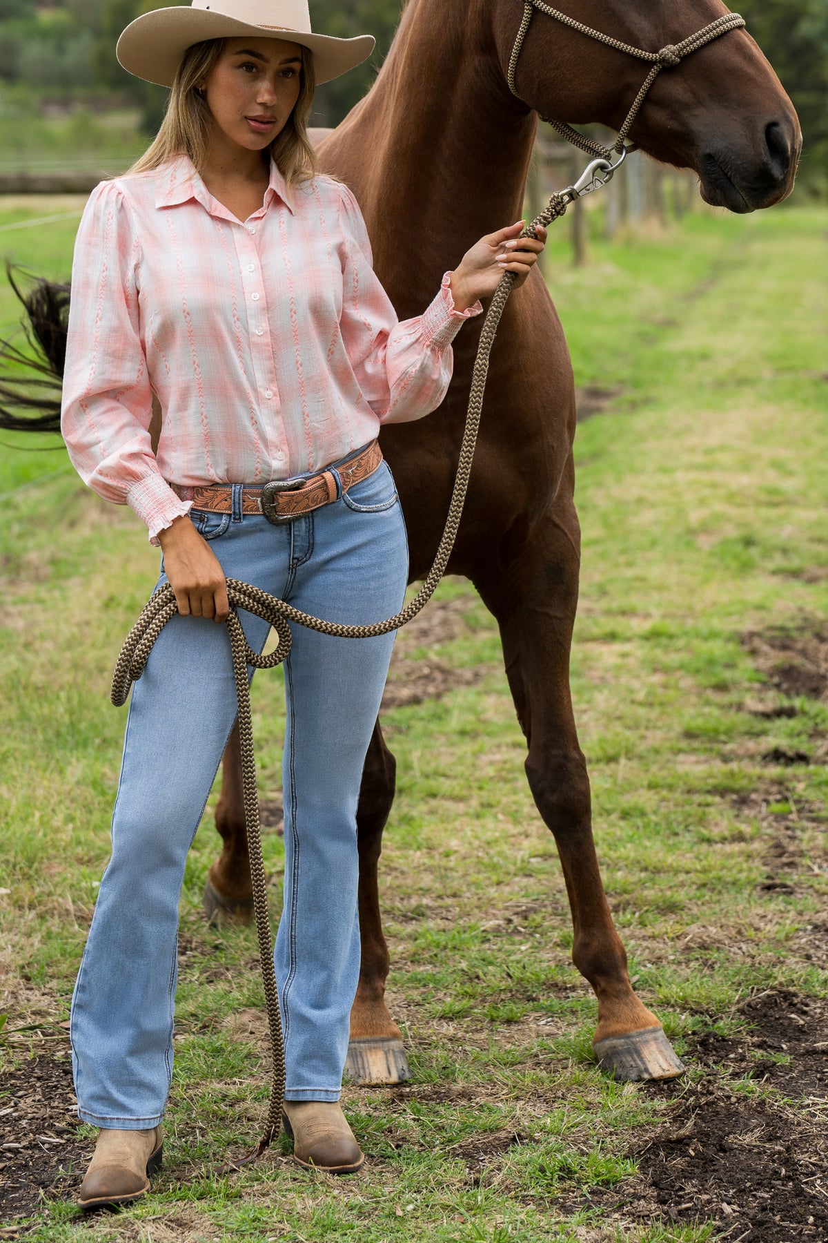 A woman wearing a light pink and white gingham long-sleeve button-up shirt, light blue jeans, a brown belt, and a beige cowboy hat stands next to a brown horse on a grassy field, holding the horse's lead rope.