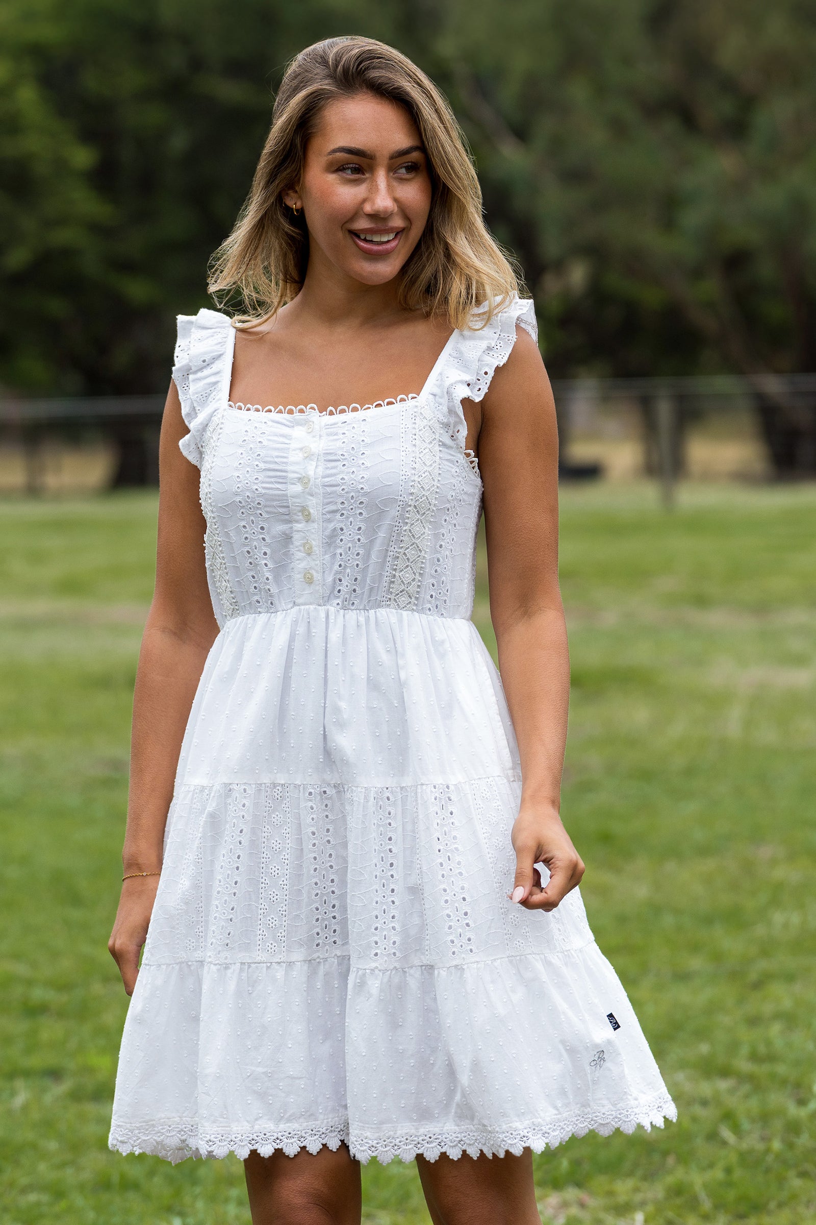 A woman wearing a white tiered eyelet dress with ruffled sleeves, standing outdoors on a grassy field with trees in the background.