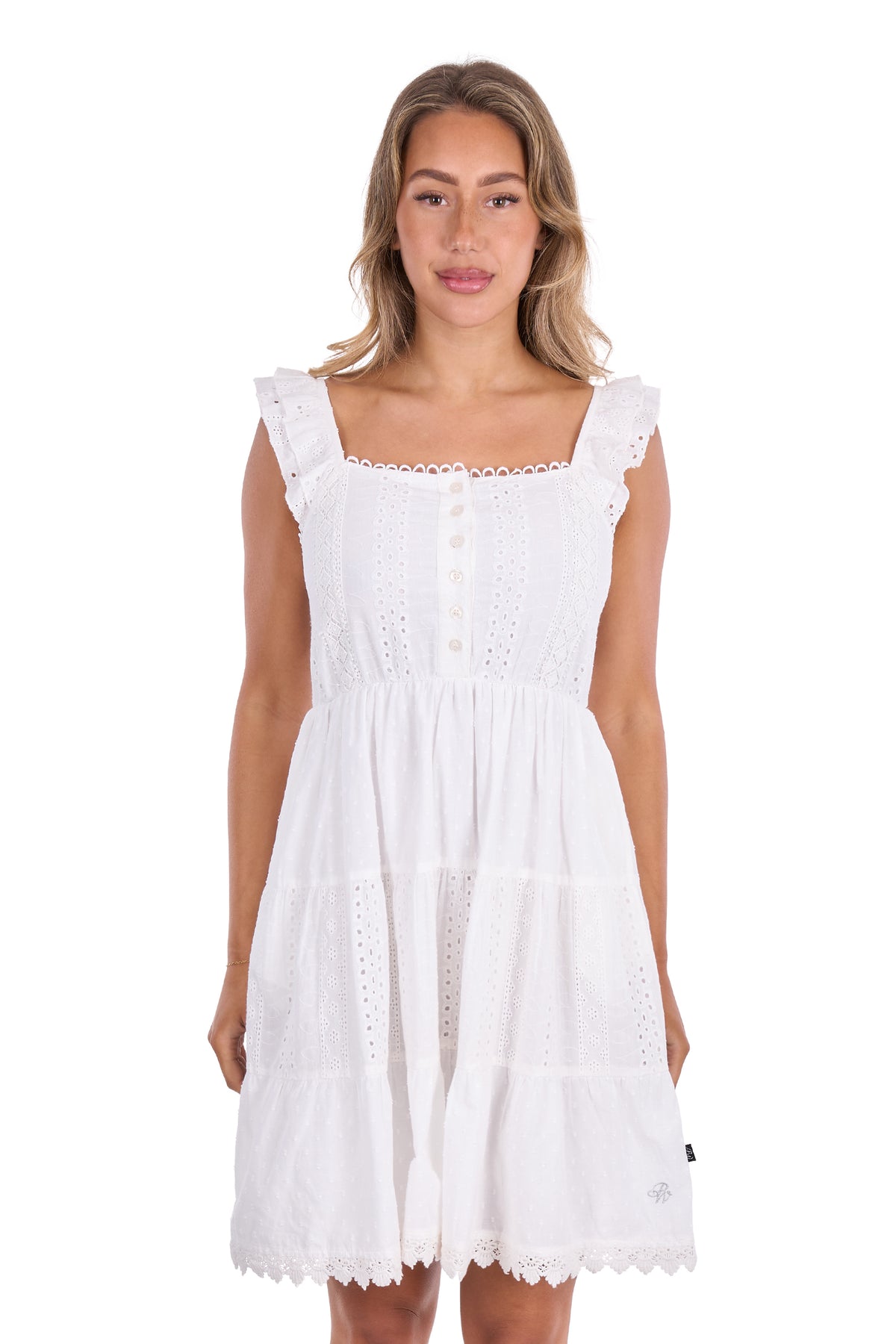 A woman modeling a white sleeveless eyelet dress with a square neckline, ruffled shoulder straps, a buttoned front, and a tiered skirt on a plain white background.