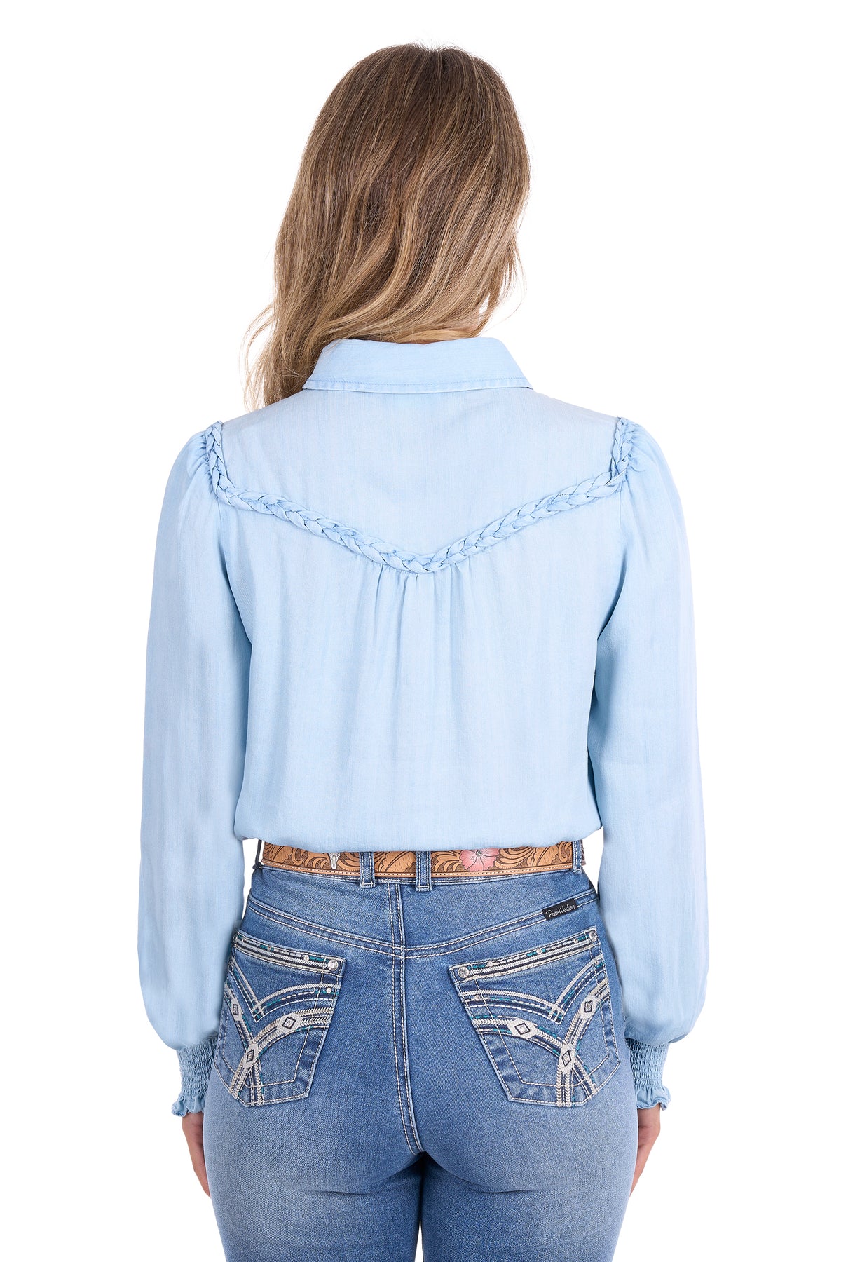 A woman shown from the back wearing a light blue long-sleeve western-style blouse with decorative braided trim across the shoulders and back, paired with blue jeans and a brown patterned belt, against a plain white background.