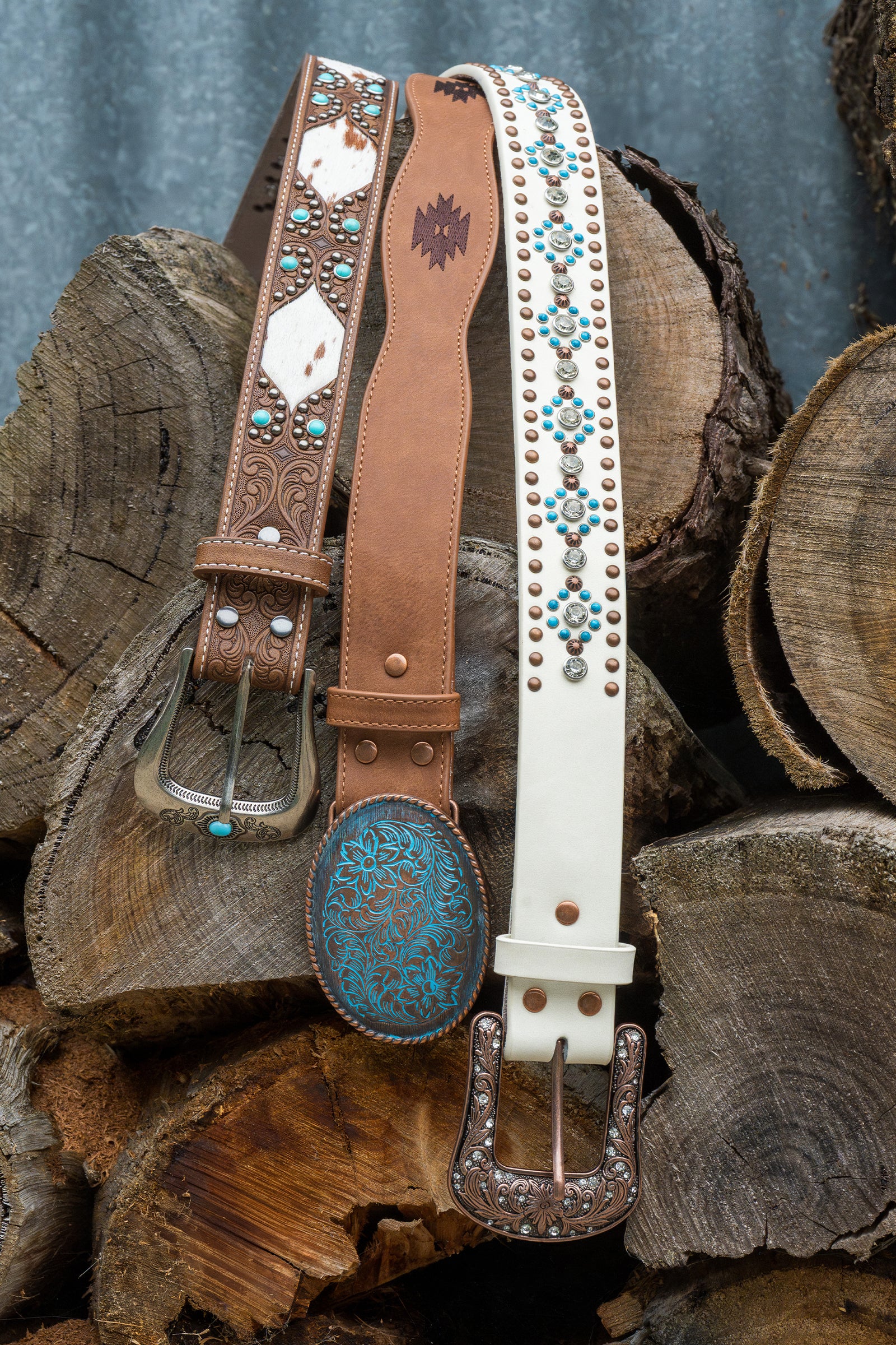 Three western-style belts displayed together on stacked wood logs: a brown tooled leather belt with turquoise and white accents, a tan leather belt with a dark brown embroidered logo, and a white leather belt with turquoise floral studs and silver studs.