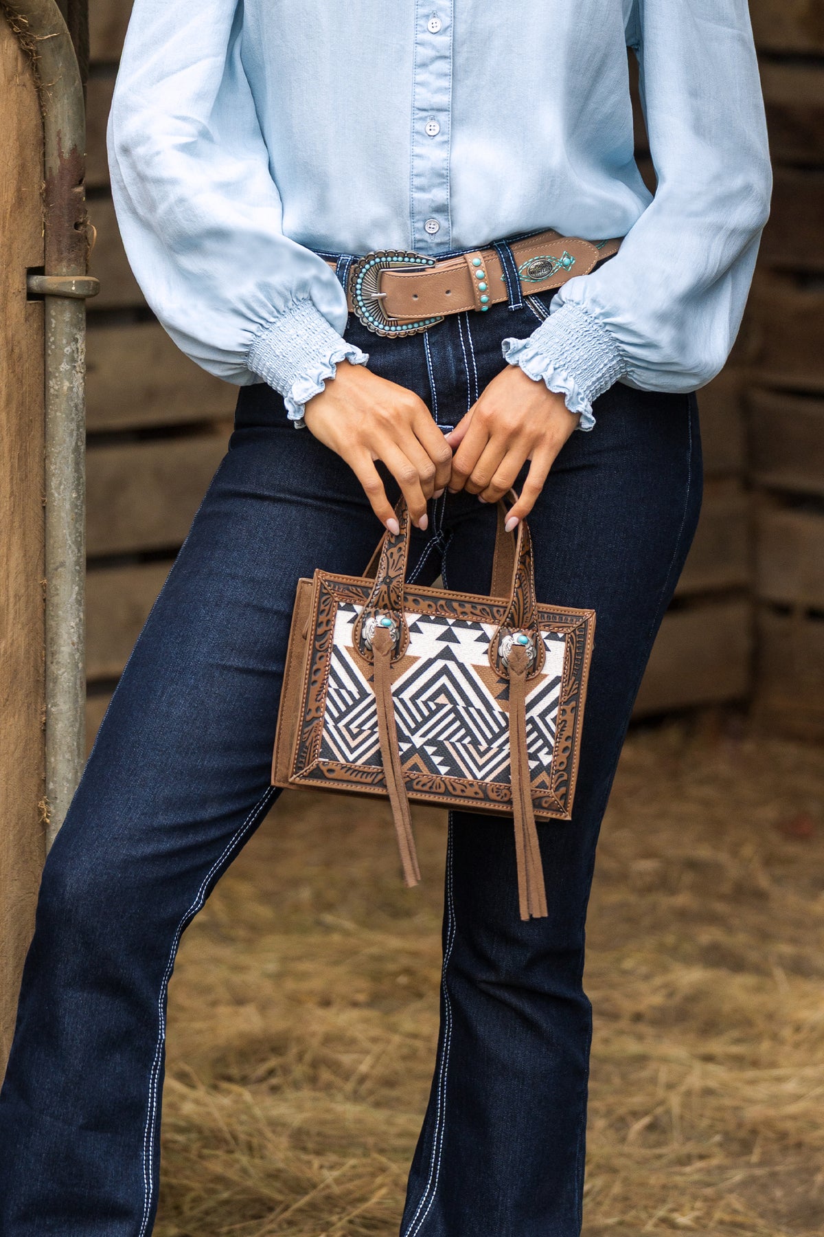 A person wearing a light blue blouse, dark blue jeans, and a western-style belt holds a brown leather handbag with a black and white geometric pattern and silver conchos.