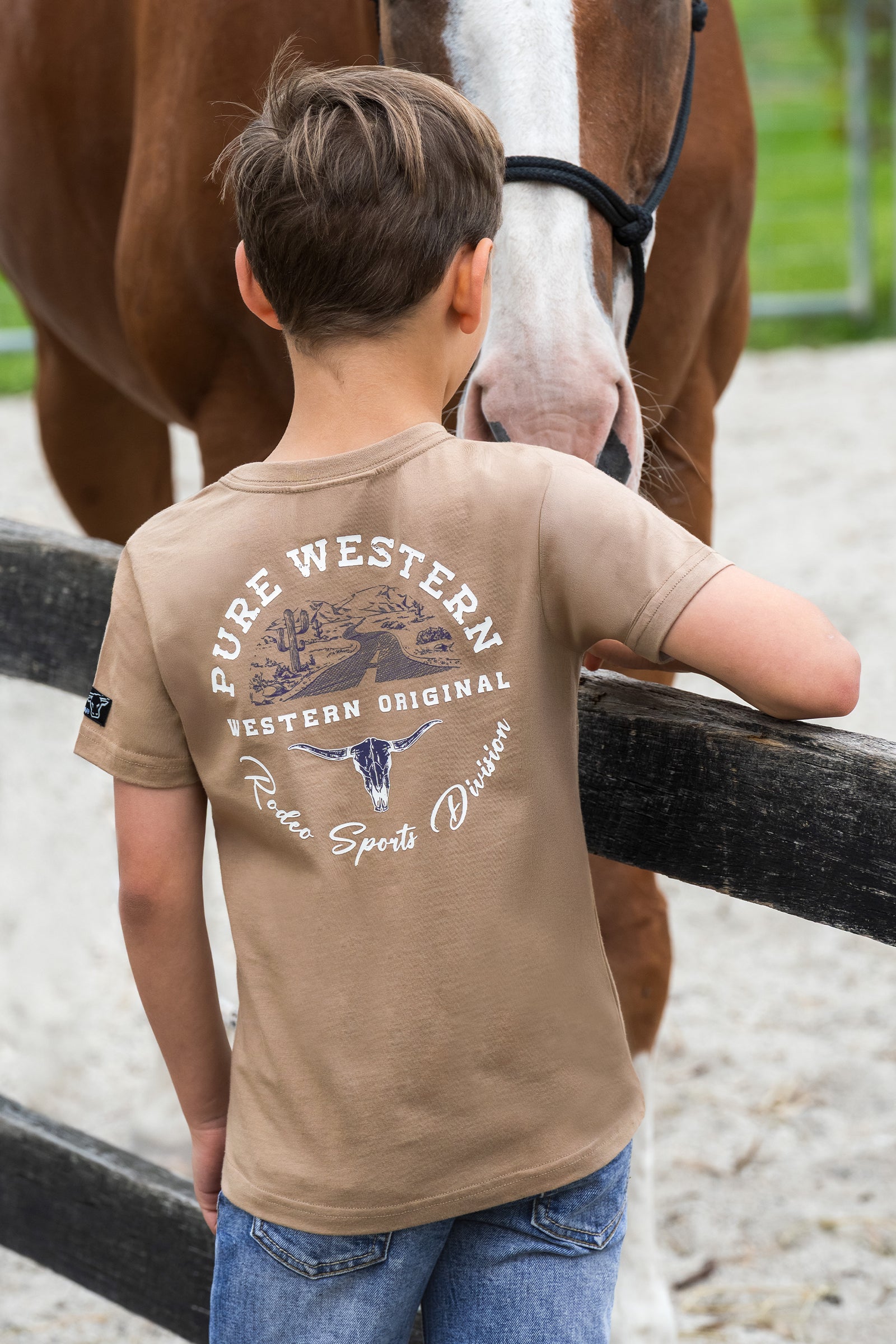 A boy wearing a tan t-shirt with a western-themed graphic on the back, standing in an equestrian arena next to a brown horse.