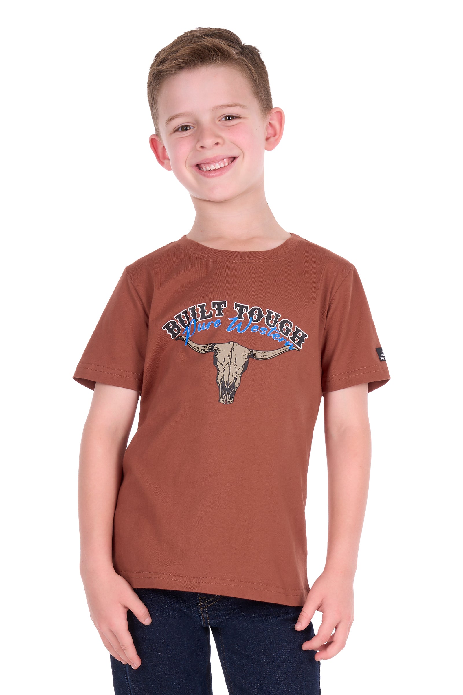 A smiling young boy wearing a rust-brown short-sleeve graphic t-shirt with a longhorn skull and 'Built Tough West Coast' text, paired with dark blue jeans, against a plain white background.