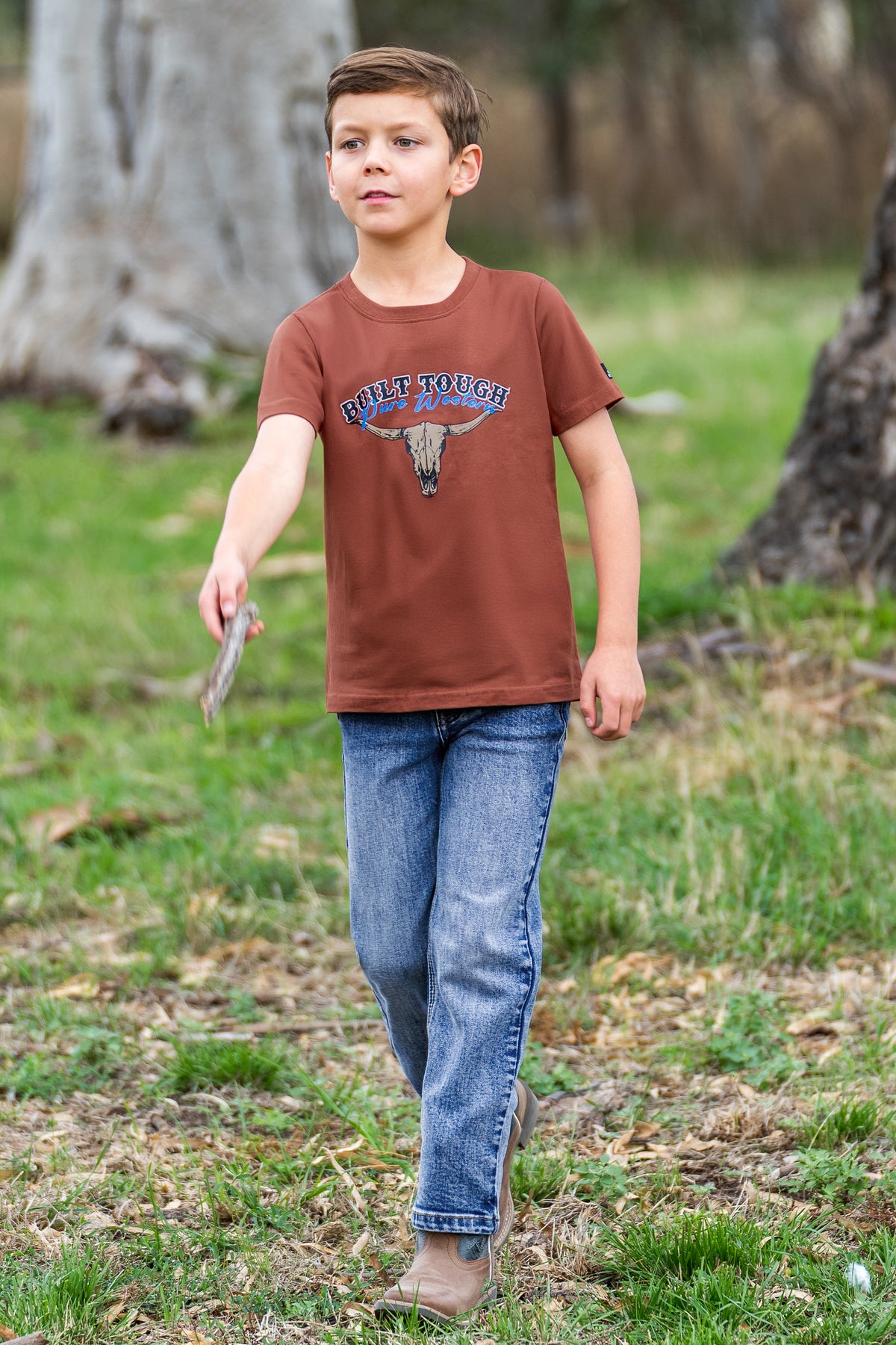 A young boy wearing a rust brown graphic t-shirt with 'RUSTIC TOUGH' and a steer skull design, paired with blue jeans, walking outdoors on a grassy path.