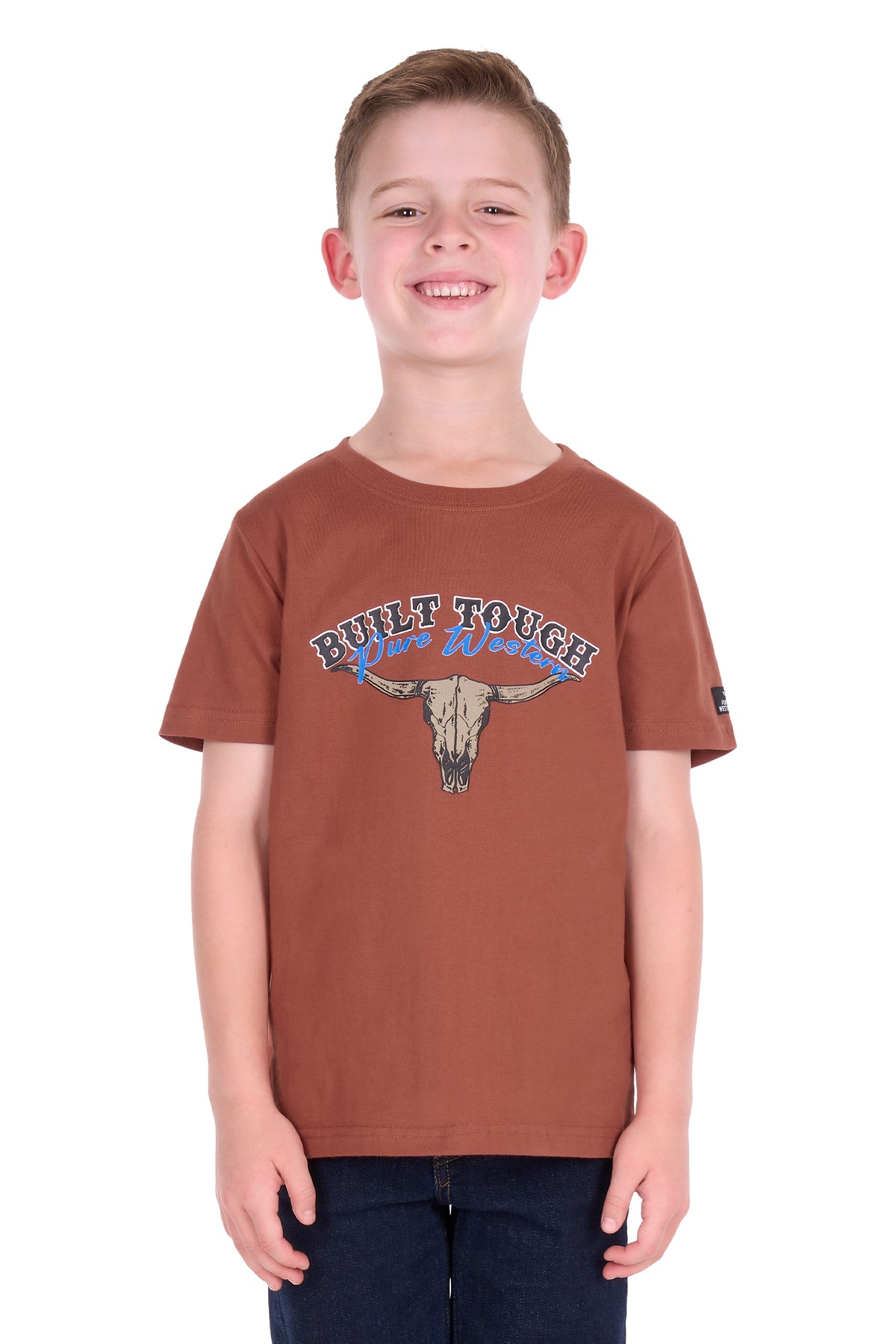 A smiling young boy wearing a rust-brown short-sleeve t-shirt with a 'Built Tough Wild West' graphic featuring a bull skull, paired with dark blue jeans, photographed against a solid white background.