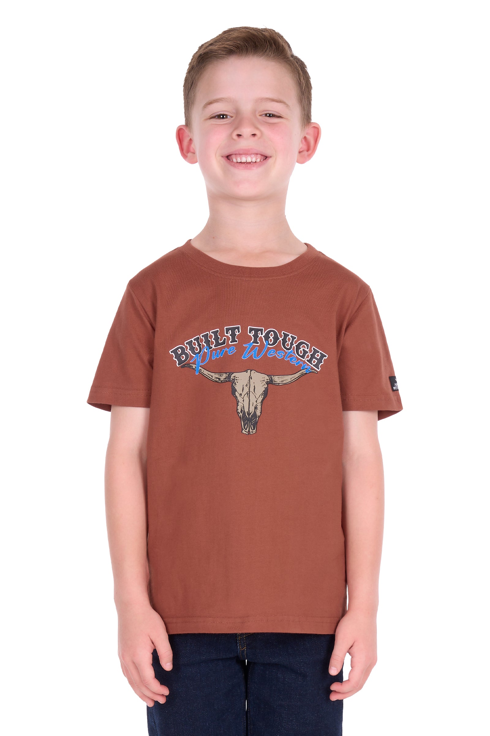 A smiling young boy wearing a rust-brown short-sleeve t-shirt with a 'Built Tough Wild West' graphic featuring a bull skull, paired with dark blue jeans, photographed against a solid white background.