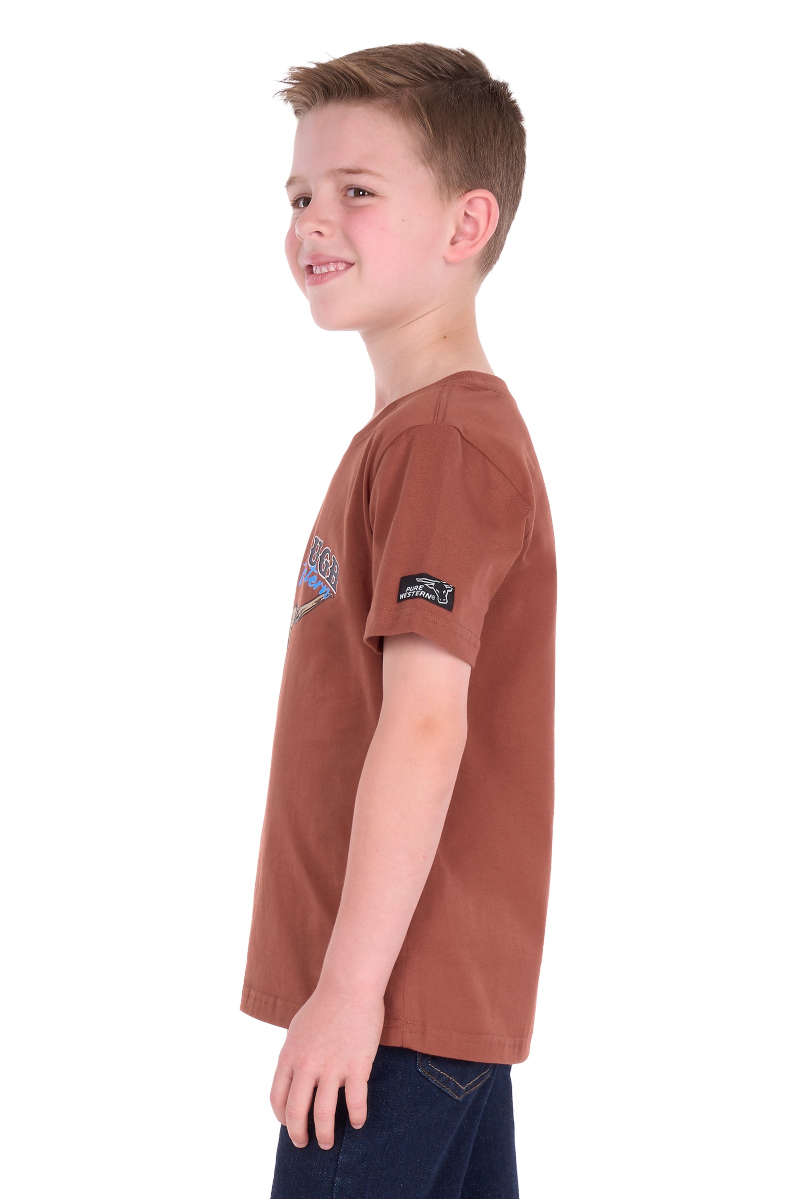 A young boy shown in profile wearing a rust-brown short-sleeve t-shirt with a small graphic on the left chest and a black brand patch on the sleeve, paired with dark blue jeans against a white background.