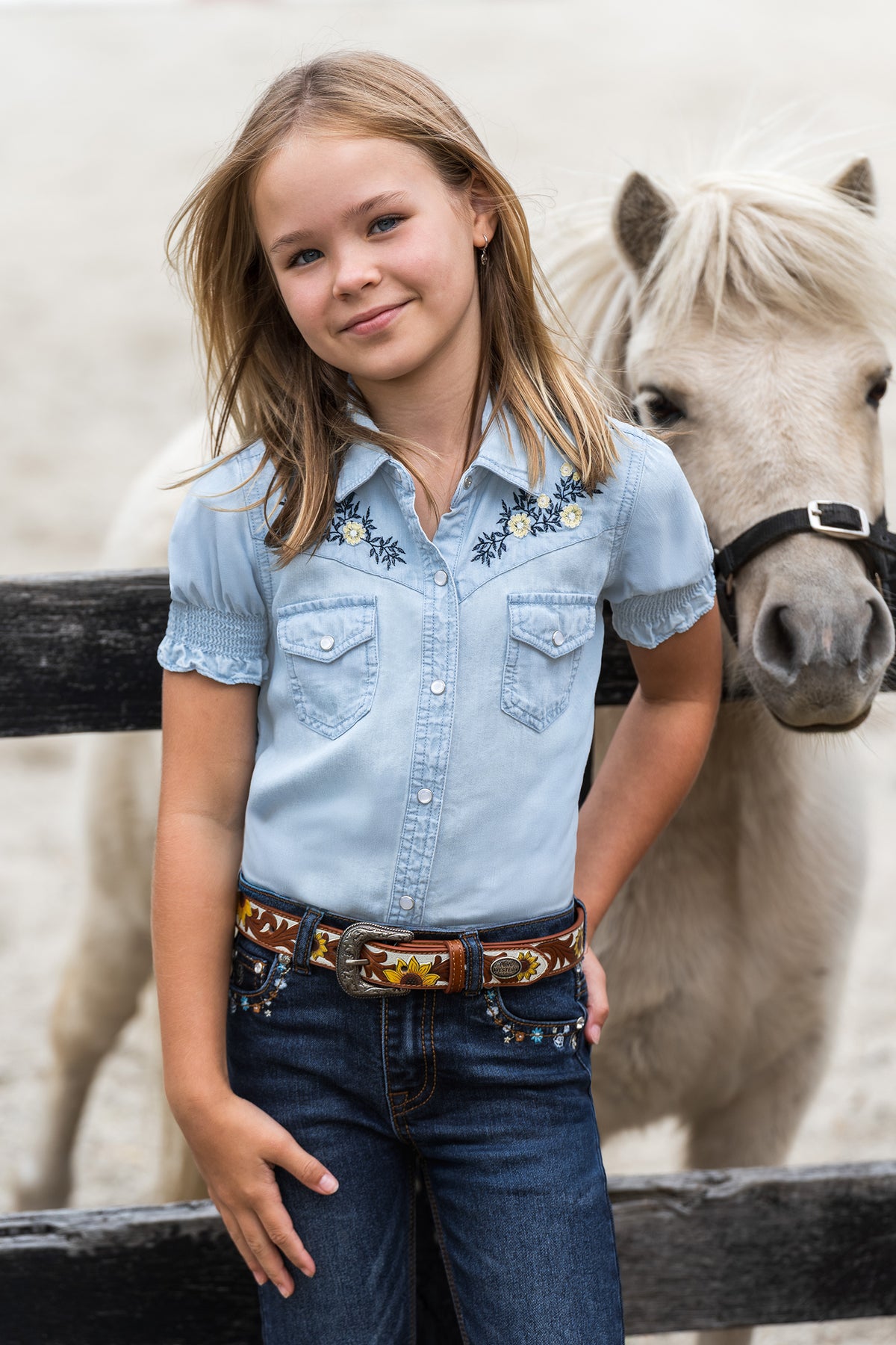 A young girl wearing a light blue western-style denim shirt with floral embroidery on the chest, tucked into dark blue jeans with a decorative brown embroidered belt, standing in front of a white pony behind a wooden fence.