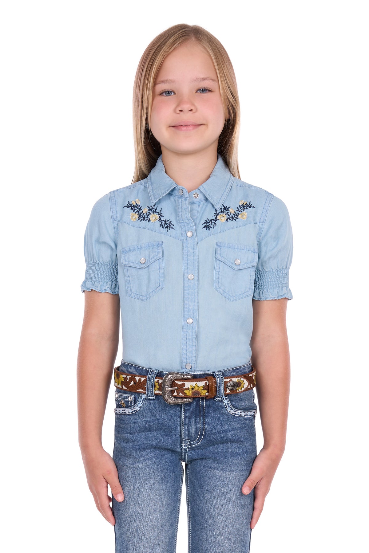A young girl with blonde hair wearing a light blue denim short-sleeve western shirt with embroidered details on the chest, paired with blue jeans and a brown western belt, against a plain white background.
