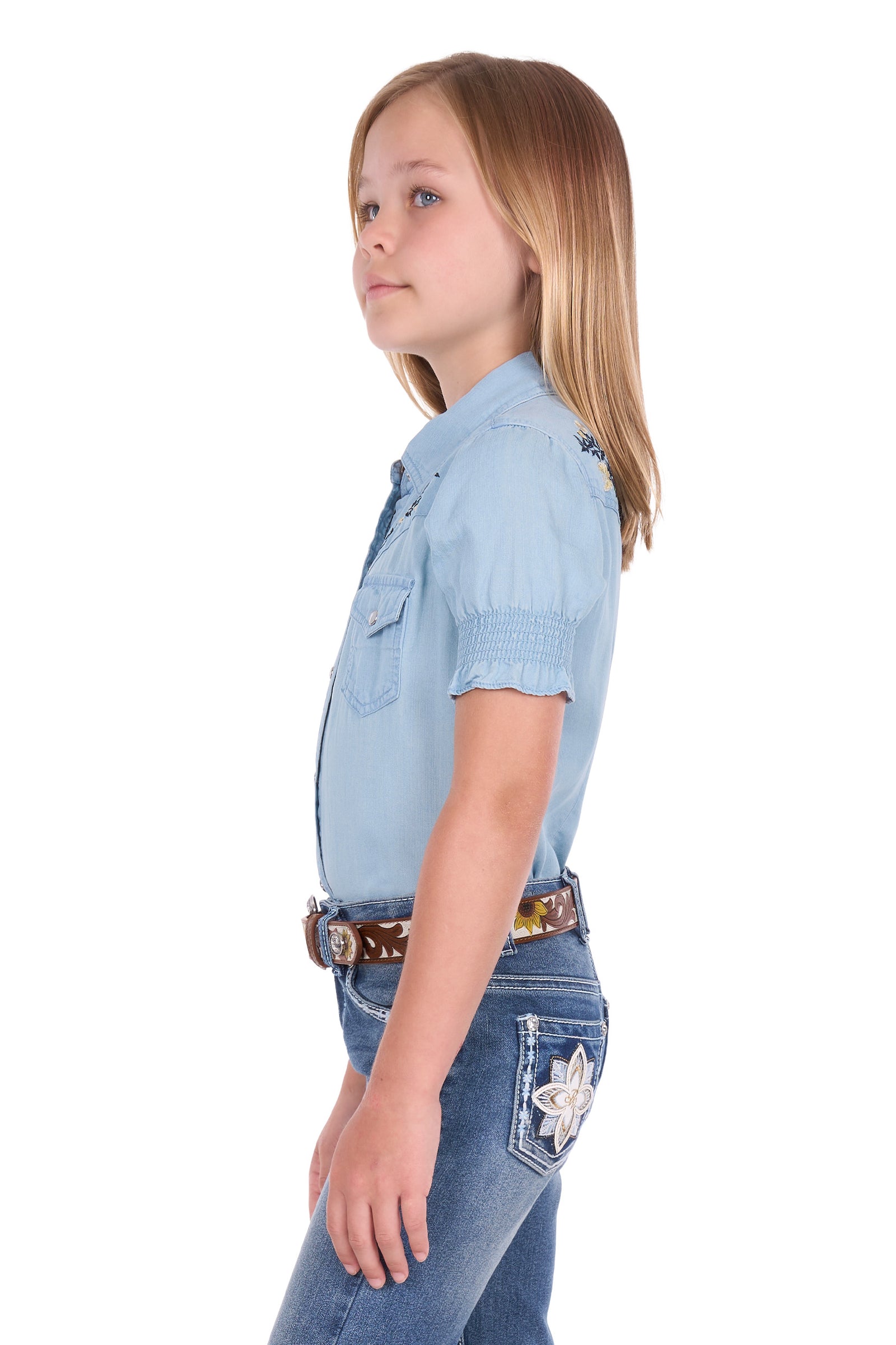 A young girl with blonde hair is shown in profile wearing a light blue short-sleeve western-style shirt, blue jeans, and a brown patterned belt against a plain white background.