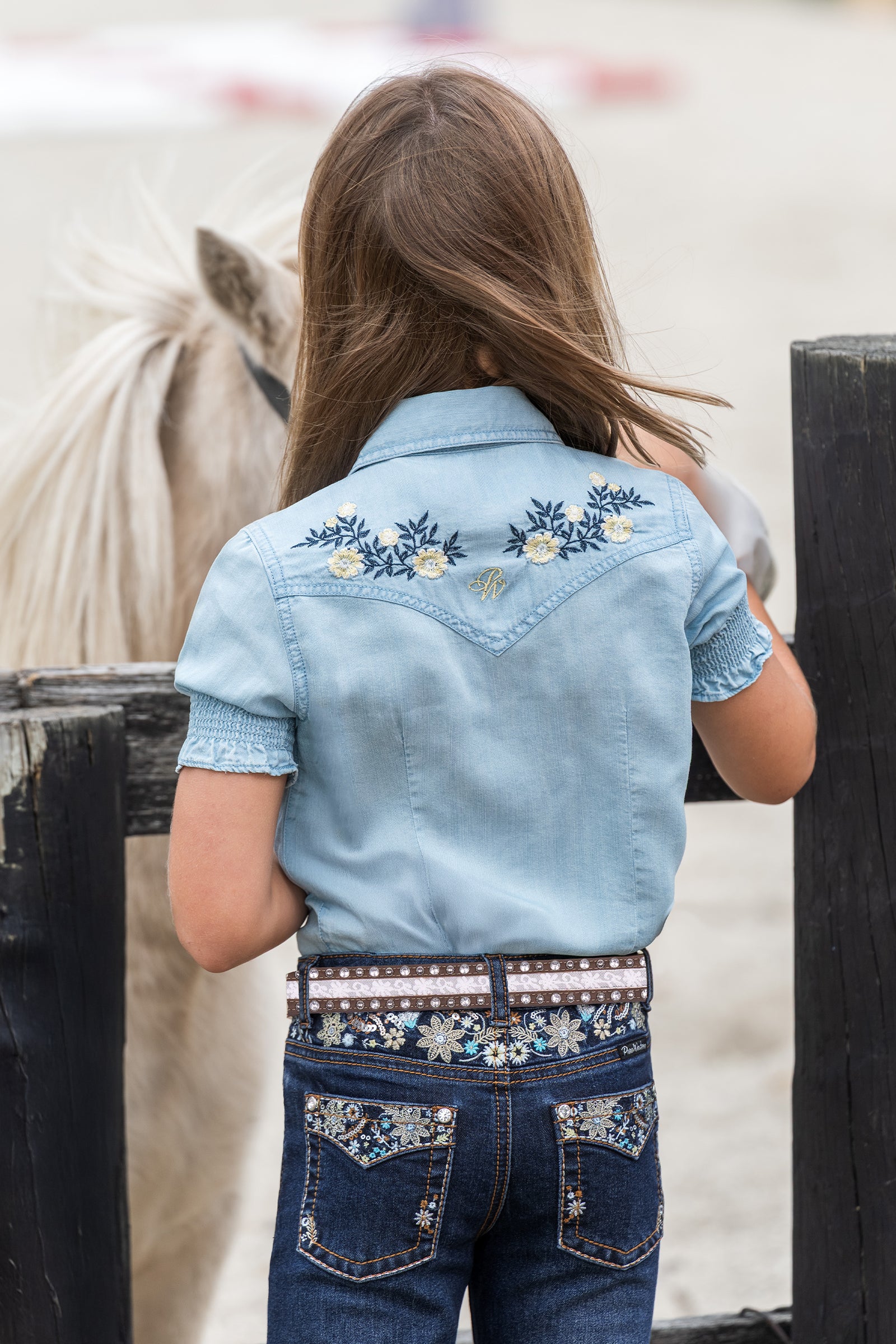 A child viewed from behind wearing a light blue western-style denim shirt with floral embroidery on the back and dark denim jeans with embroidered pocket details and a decorative belt, standing near a horse fence in an outdoor setting.