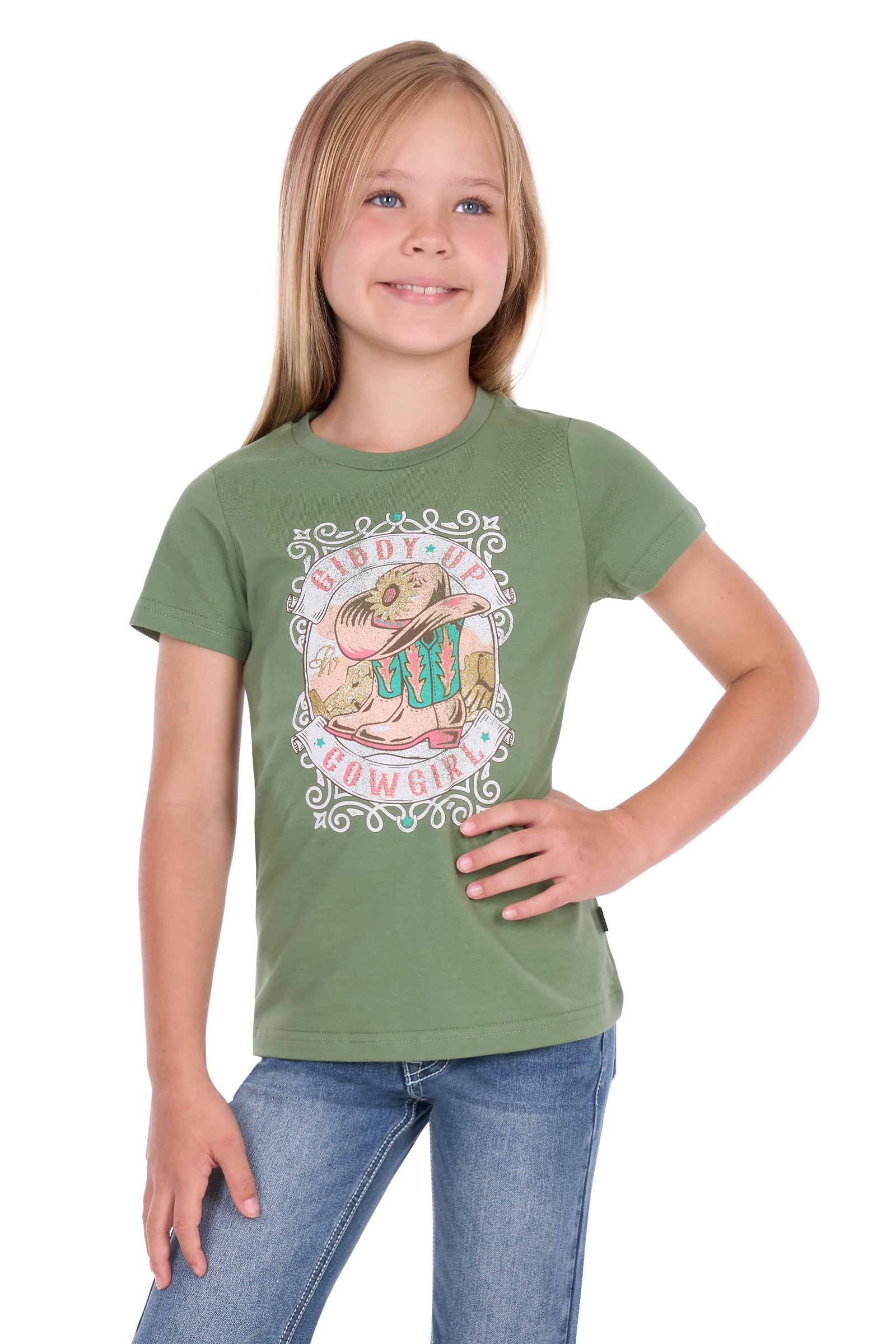 A young girl with blonde hair wearing an olive green short-sleeve t-shirt with a cowgirl graphic and blue jeans, standing against a white background.