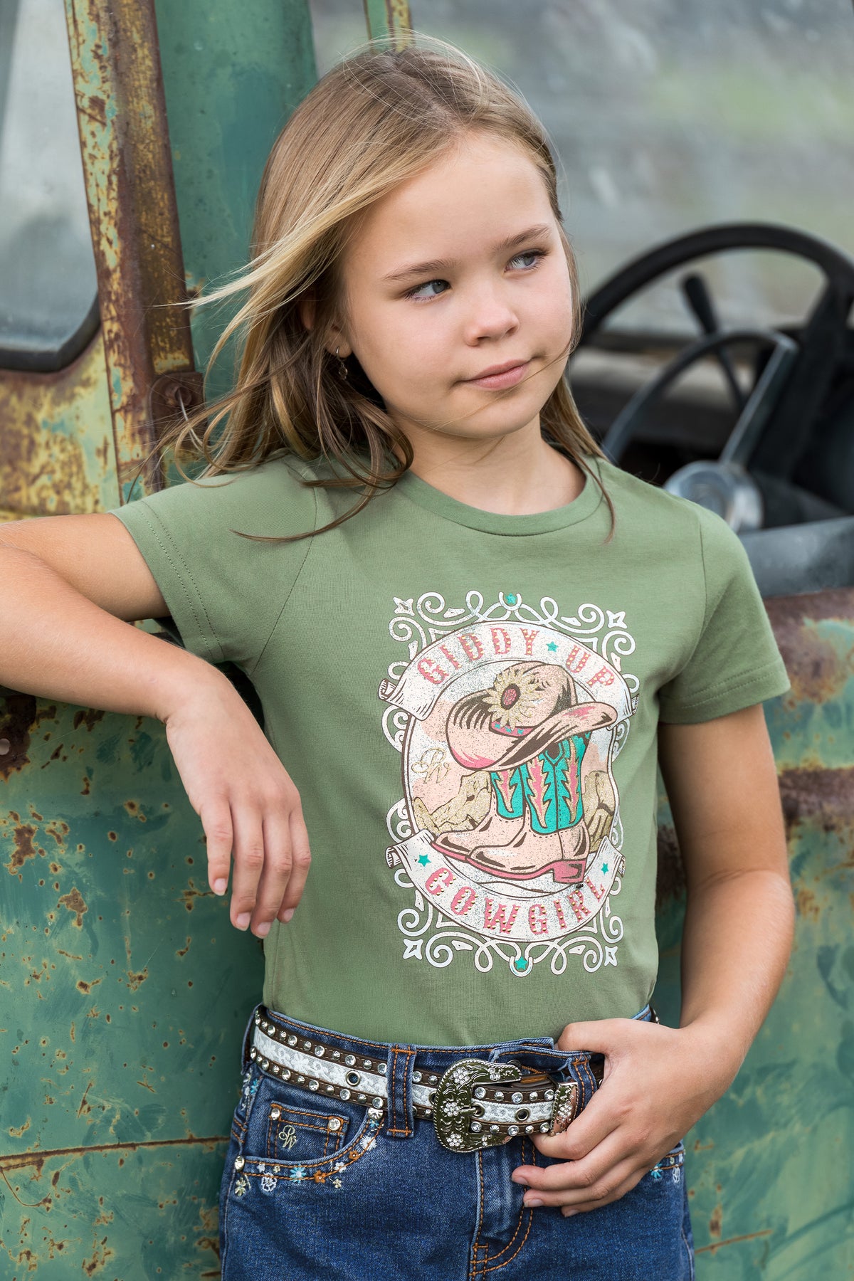 A young girl with blonde hair wearing an olive green graphic t-shirt and a studded denim belt, posing in front of a weathered green truck.