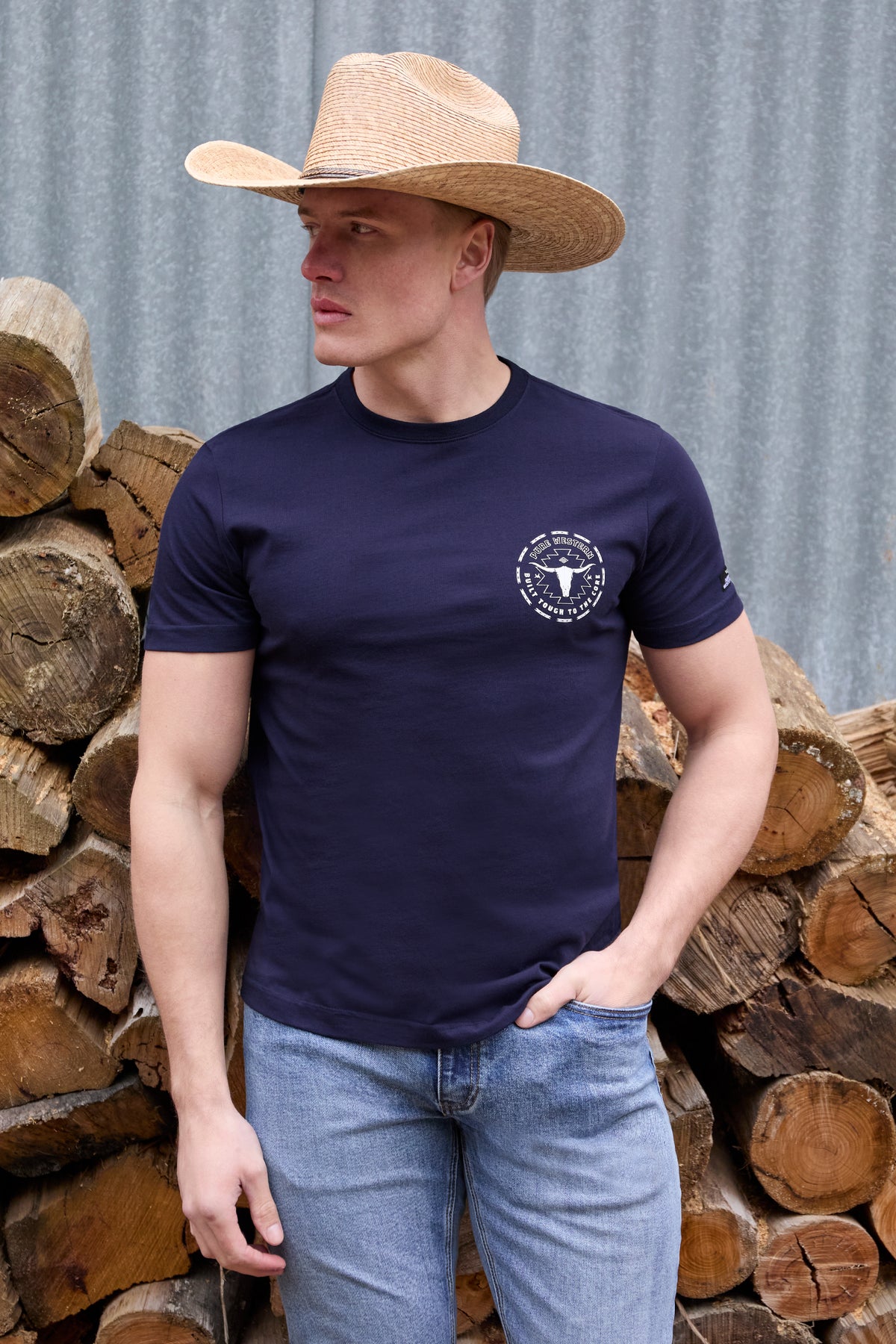 Man wearing a navy t-shirt with a logo and a cowboy hat, standing in front of stacked logs.