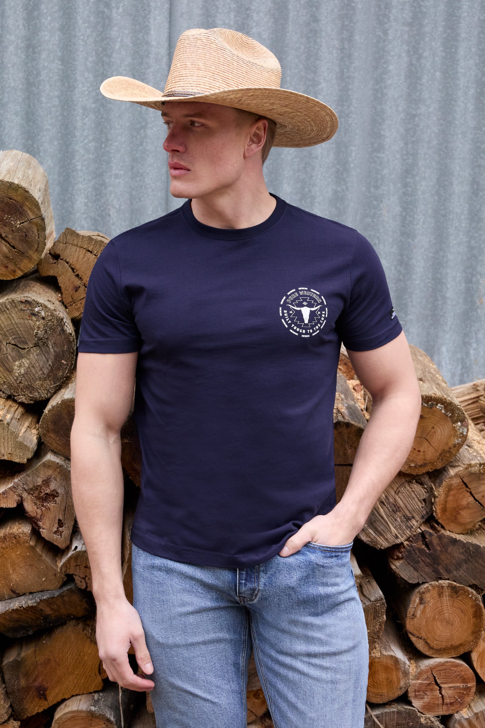 Man wearing a navy t-shirt with a logo and a cowboy hat, standing in front of stacked logs.