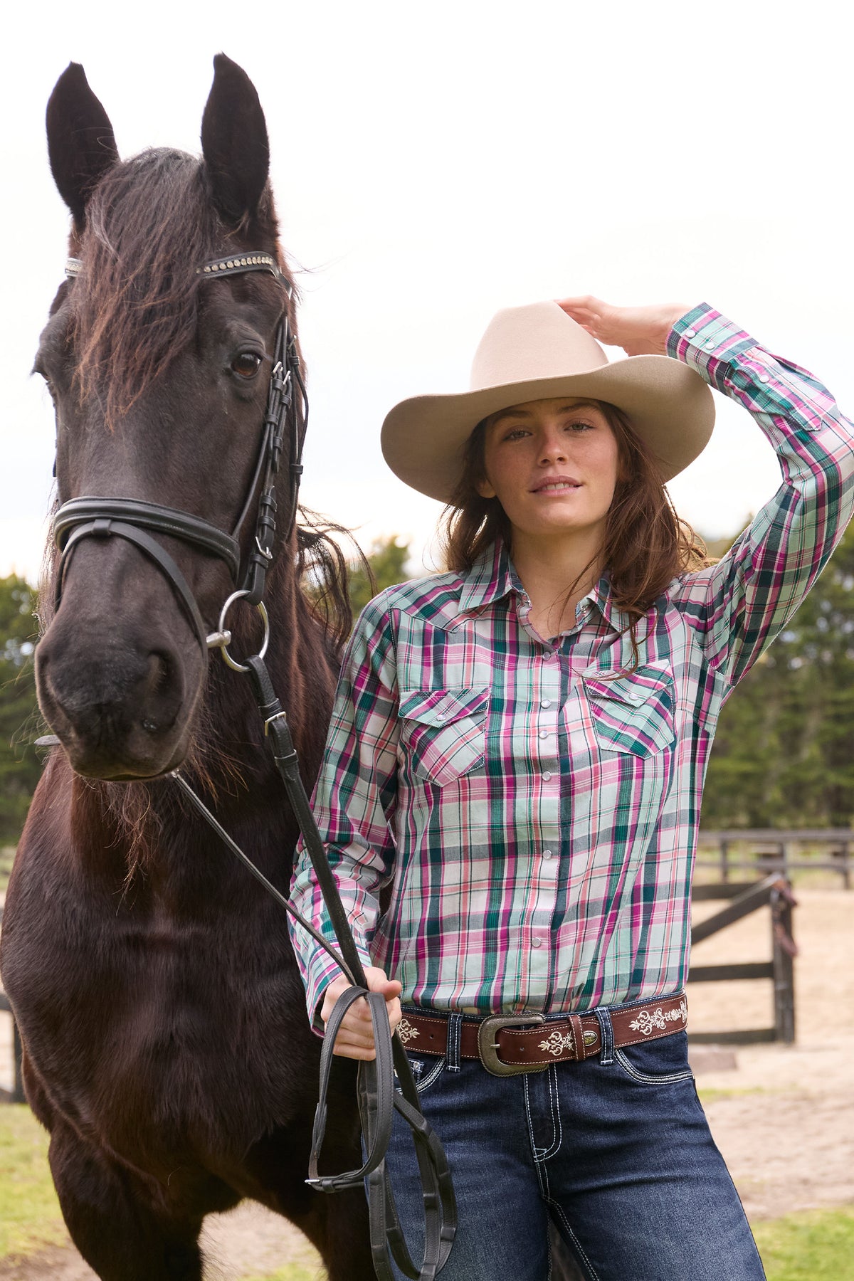 Woman in plaid shirt and cowboy hat standing next to a horse outdoors.