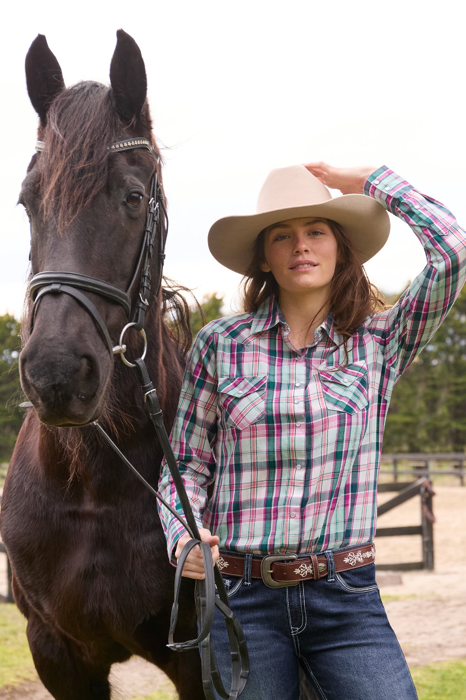 Woman in plaid shirt and cowboy hat standing next to a horse outdoors.