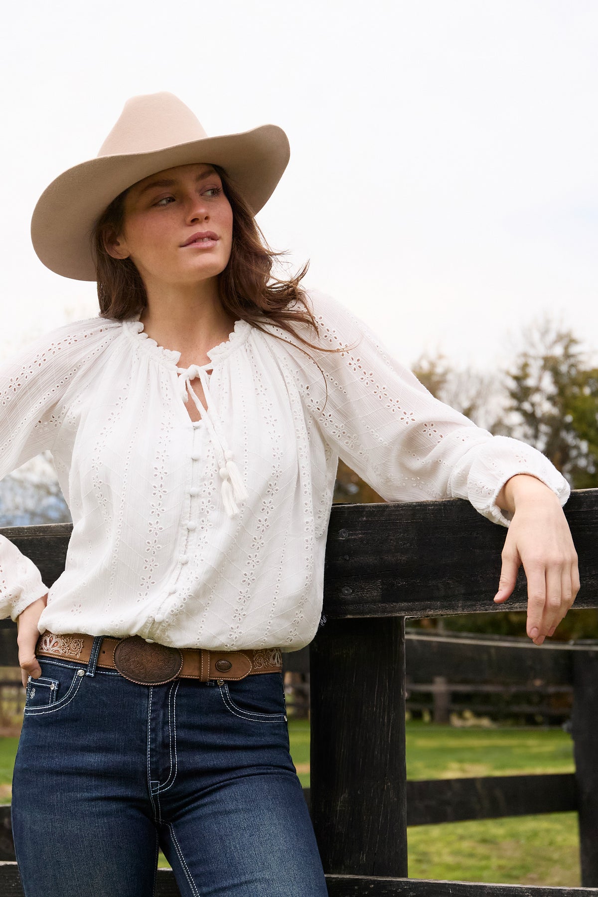 Woman wearing a white blouse, blue jeans, and a beige hat, standing by a wooden fence.