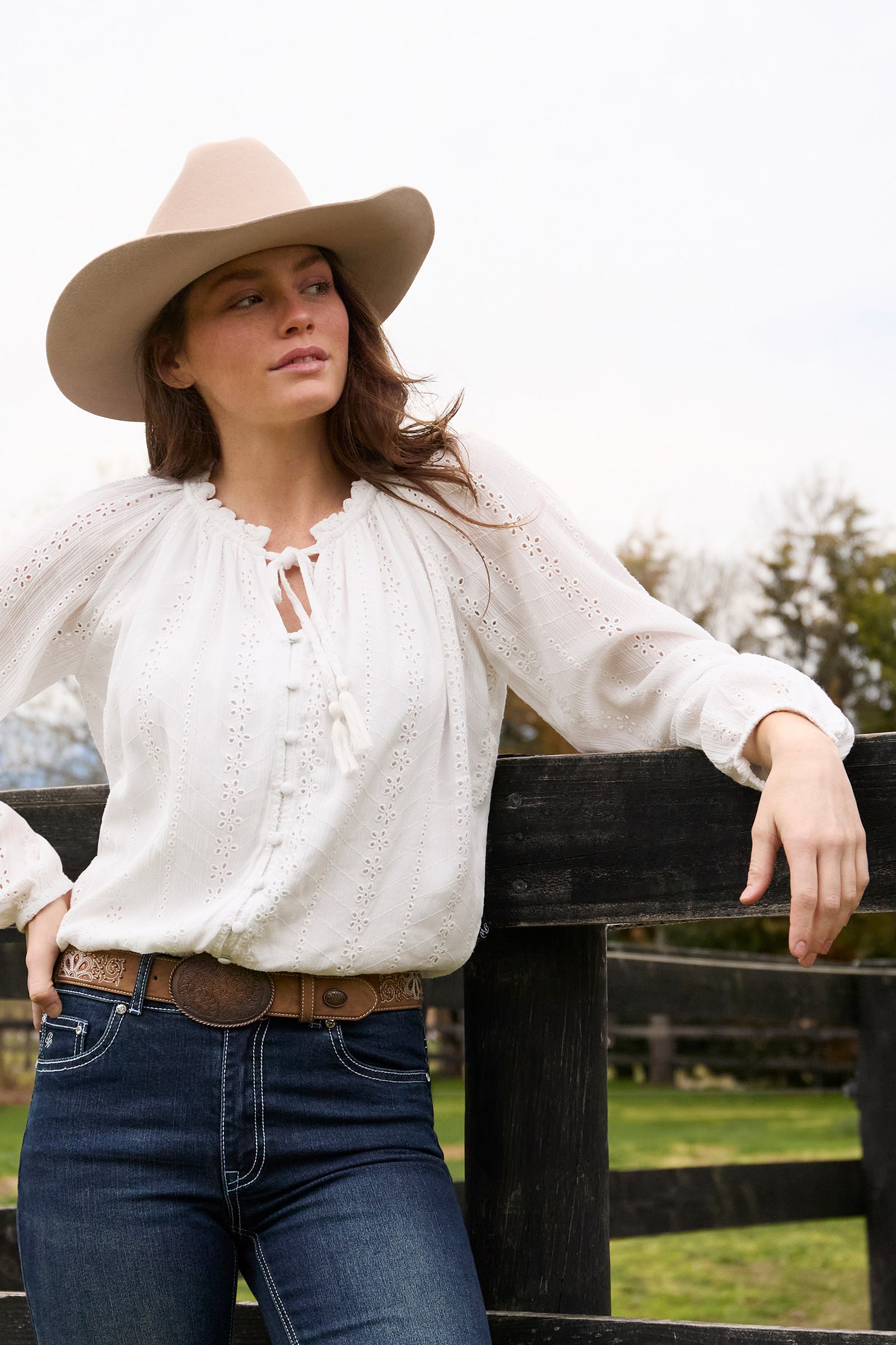 Woman wearing a white blouse, blue jeans, and a beige hat, standing by a wooden fence.