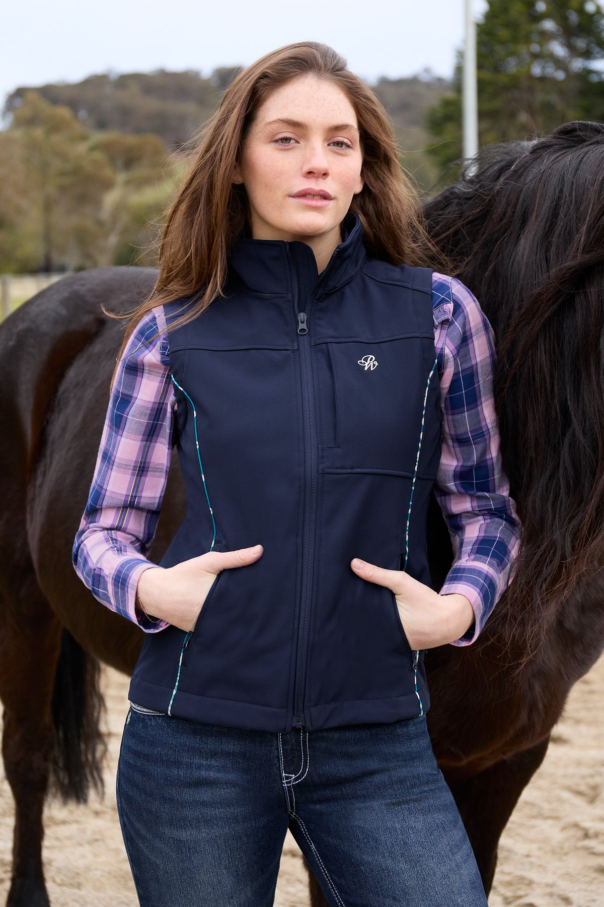 Woman wearing a navy vest with a logo, standing next to a horse in an outdoor setting.