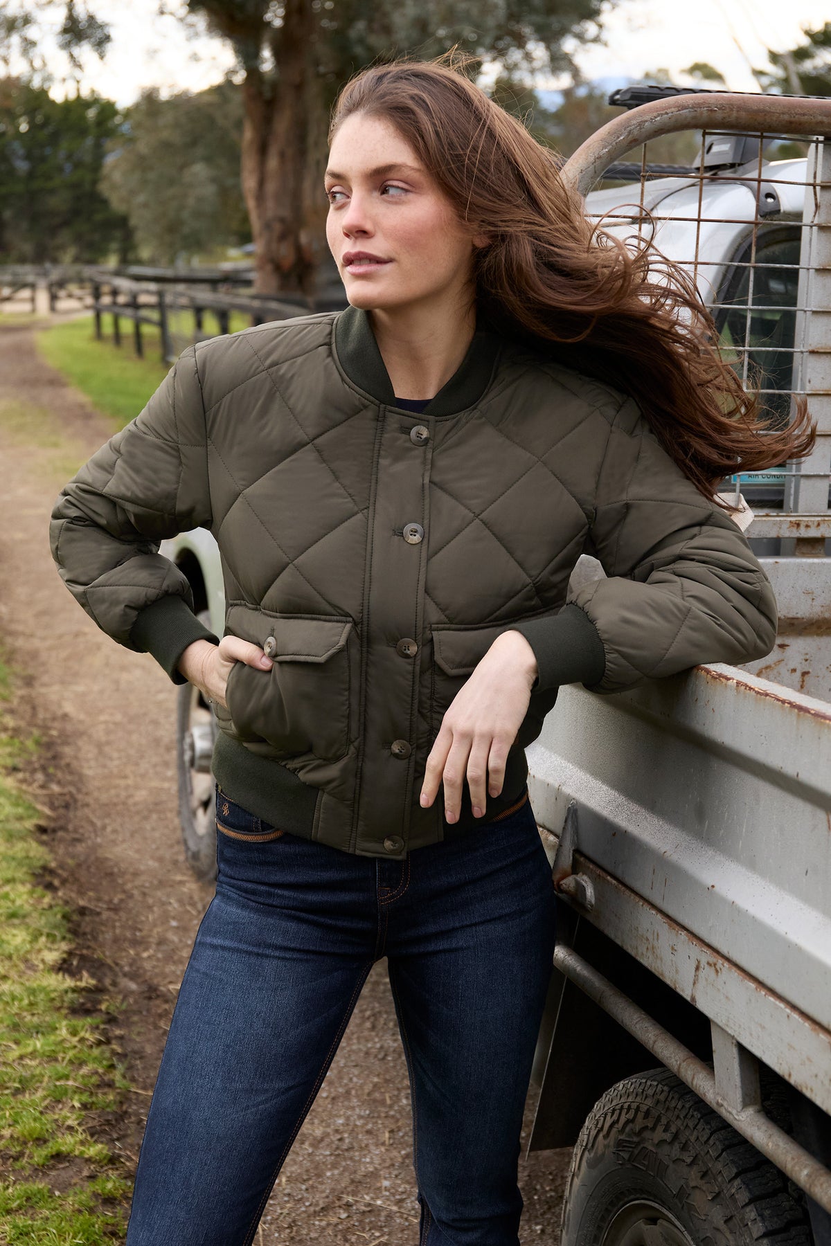 Woman wearing a quilted jacket standing next to a truck on a dirt road.