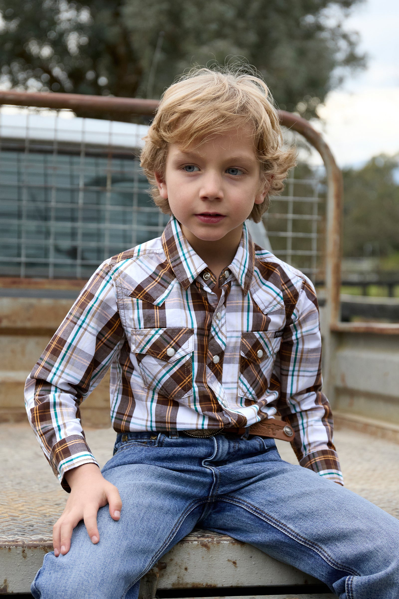 Young boy wearing a plaid shirt and jeans sitting on a metal structure outdoors.
